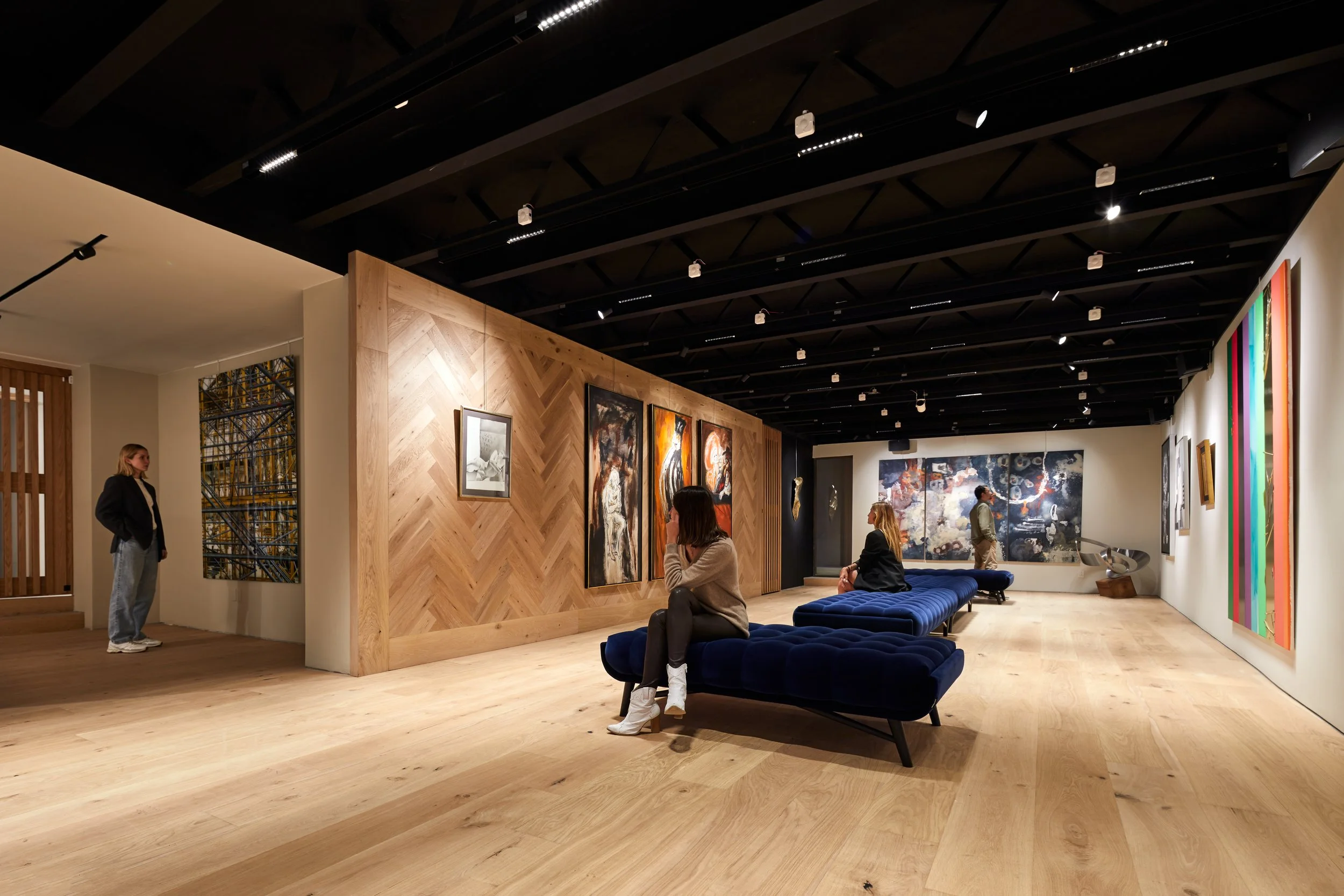 Vista interior de una galería de arte con varias obras en las paredes, bancos azules y personas observando los cuadros.
