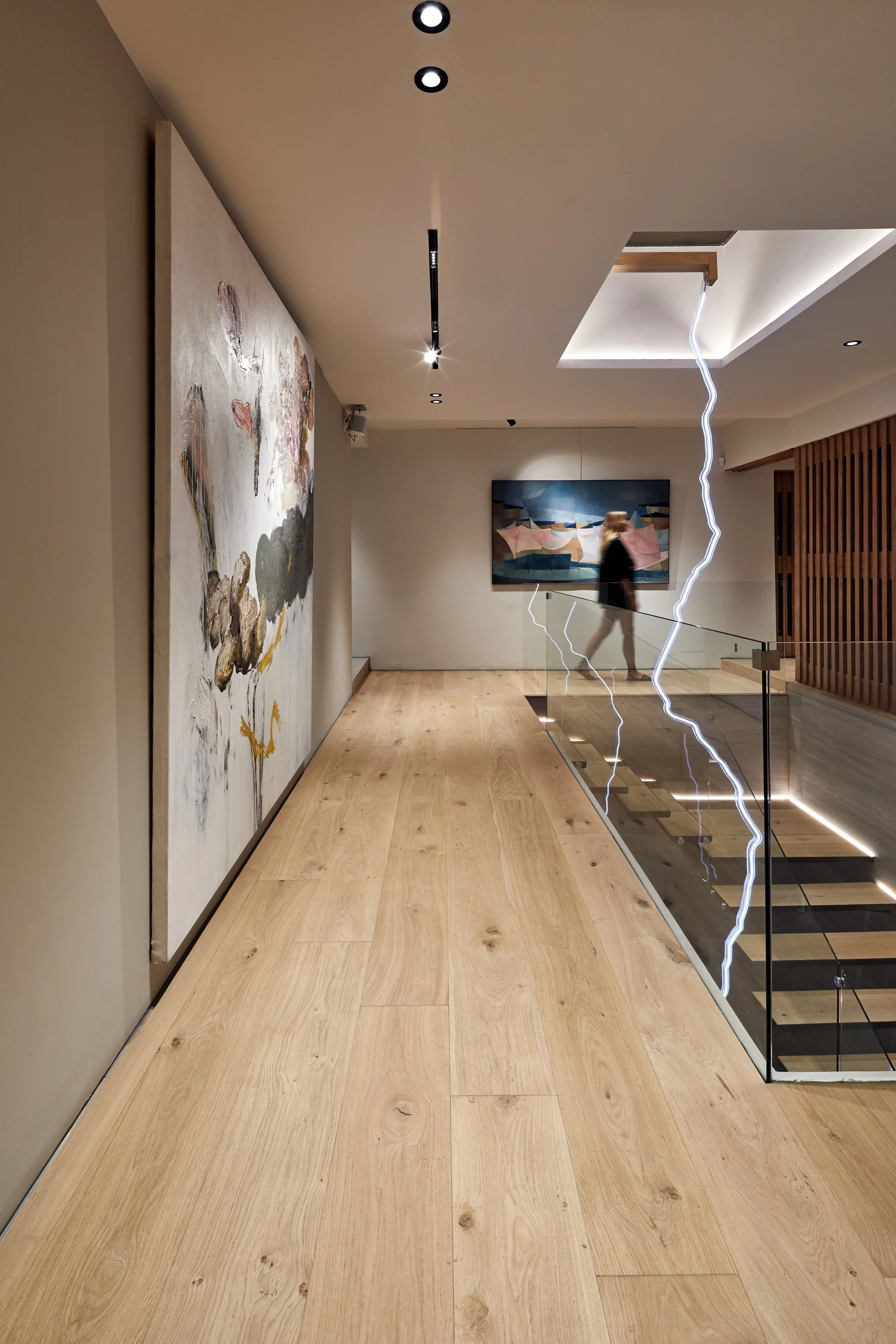 Interior de una galería de arte con pinturas en las paredes, iluminación moderna en el techo y un sótano visible por una barandilla de vidrio que tiene relámpagos eléctricos corriendo a través de ella, y una persona caminando en el fondo.