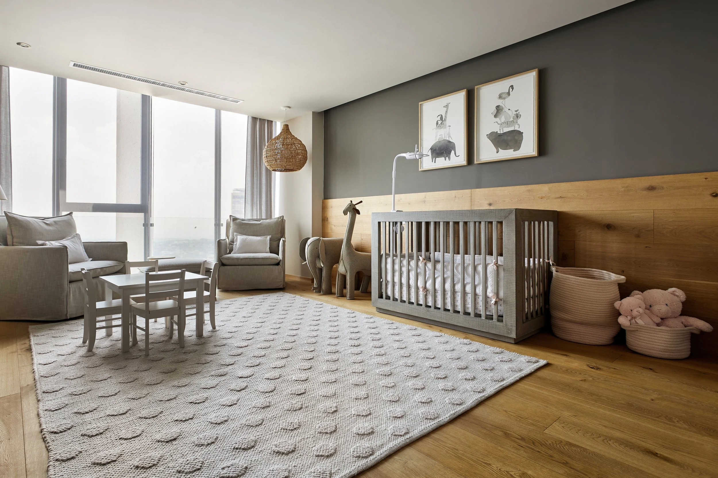 Sala de estar con sillones, una alfombra blanca con detalles en relieve, ventanas grandes, decoración infantil con jirafas y arte en la pared, cuna, cestas con peluches y una lámpara de techo de fibra natural.