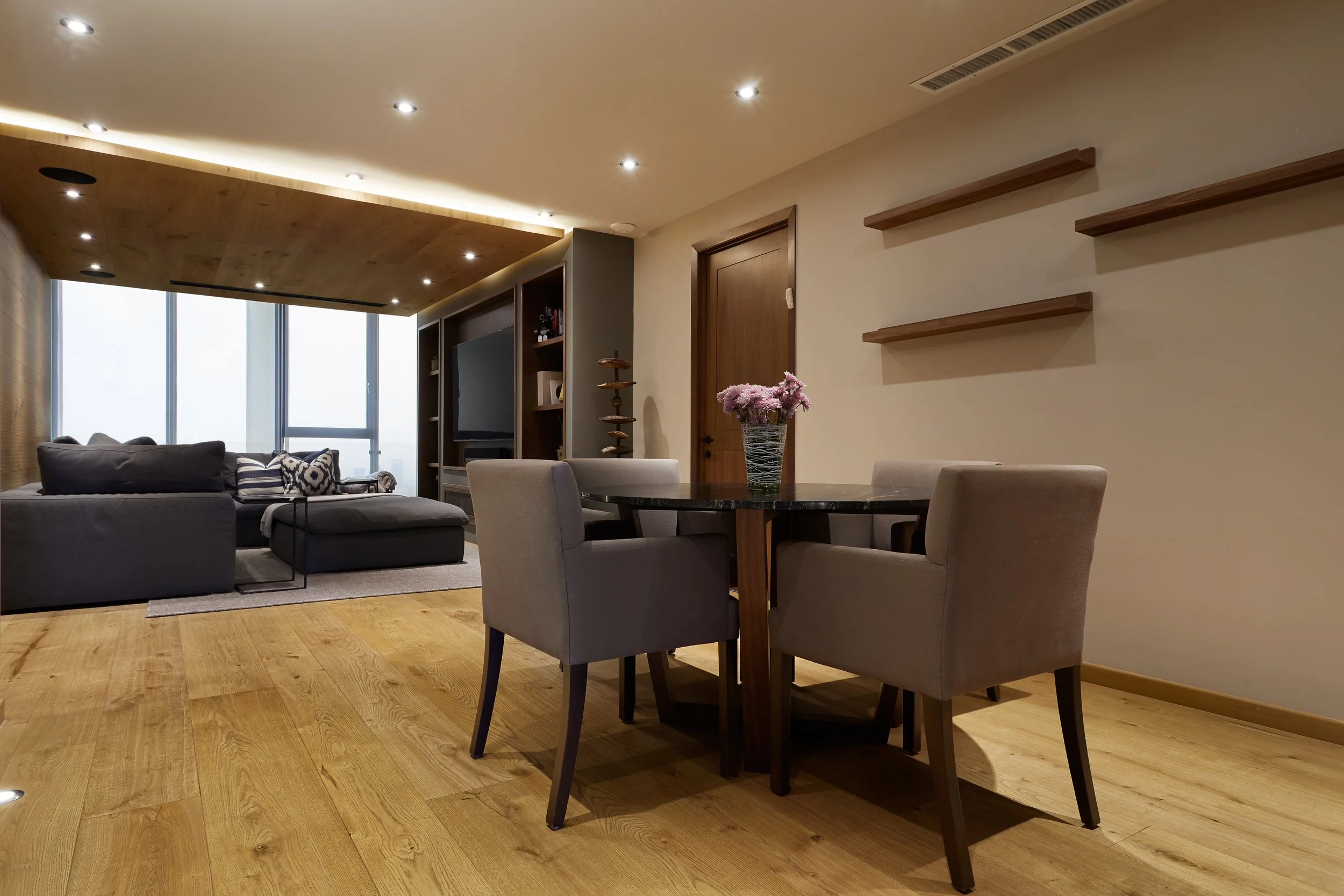 Sala de estar y comedor con piso de madera, sofá gris, mesa de comedor con cinco sillas beige y jarrón de flores rosas en la mesa.