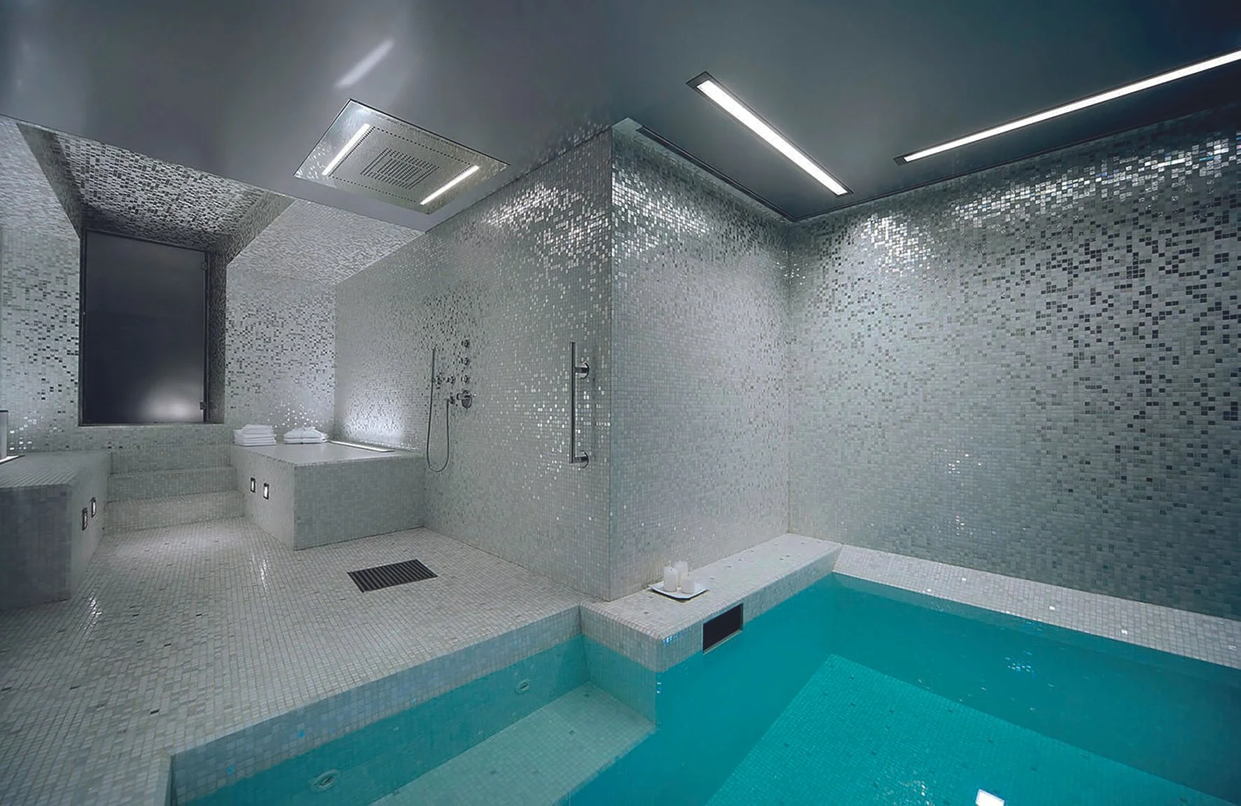 Piscina y área de baño con paredes y pisos de azulejos plateados y una pequeña piscina con agua azul.