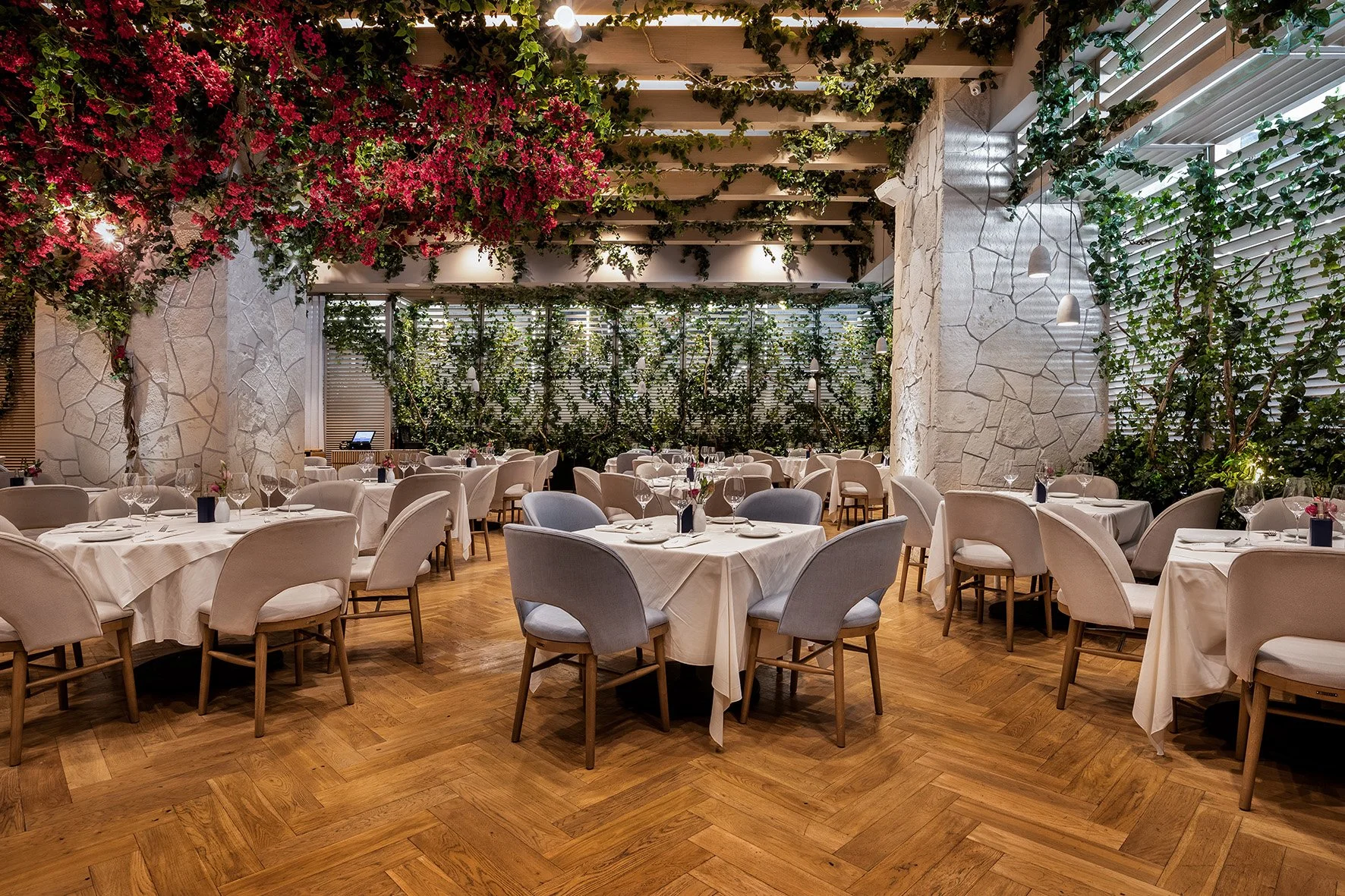 Comedor elegante con mesas vestidas con manteles blancos, sillas acolchonadas y copas de vino, decorado con plantas y flores, con paredes de piedra y paredes de vidrio con persianas