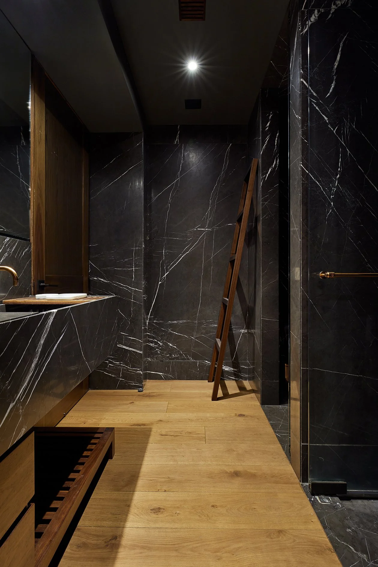 Interior de baño con paredes de mármol negro con vetas blancas, suelo de madera, espejo y escalera de madera.