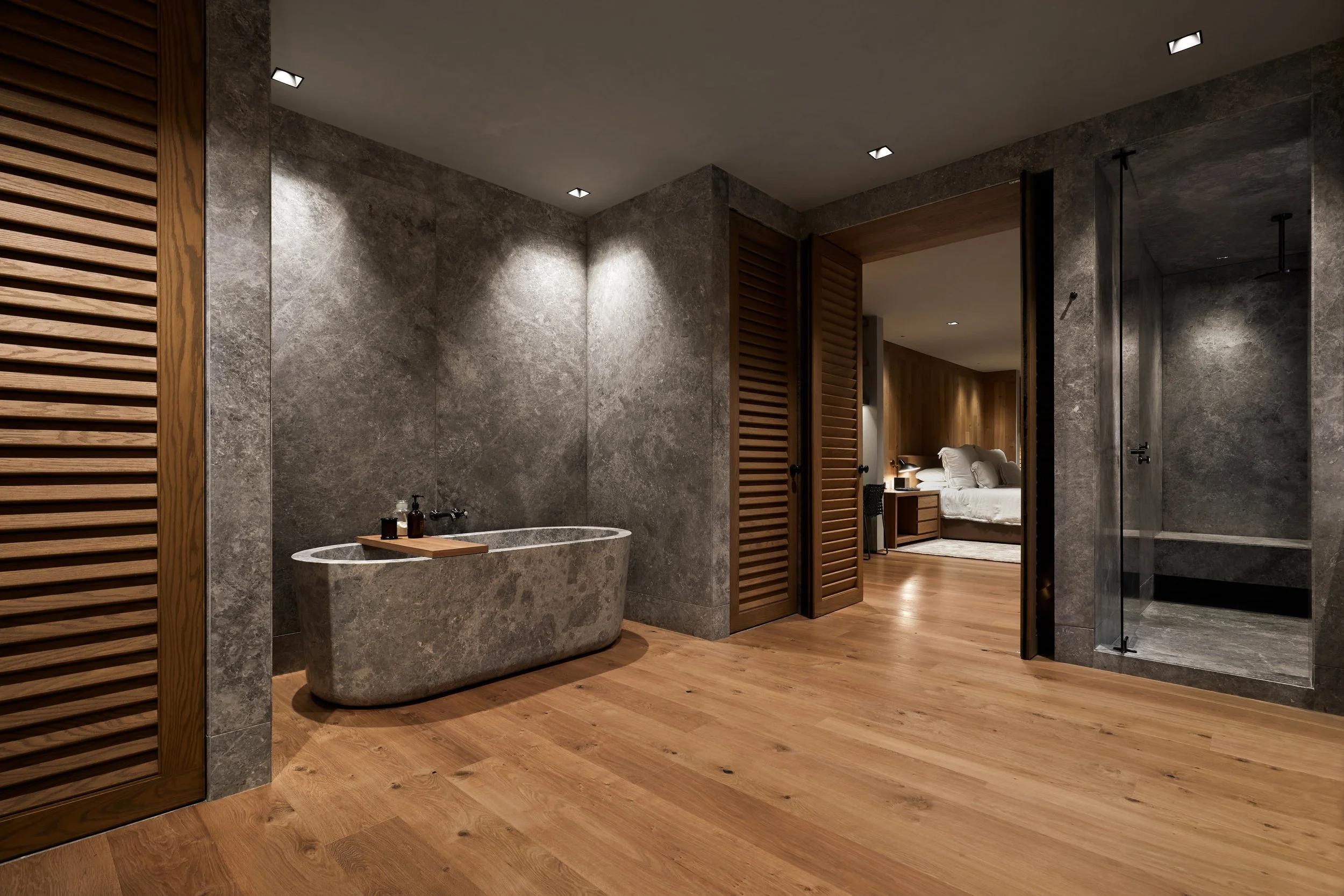 Baño con paredes de piedra gris, ducha con puerta de cristal y bañera de piedra, piso de madera y puerta de lamas de madera que lleva a una habitación con cama y mesitas de noche.