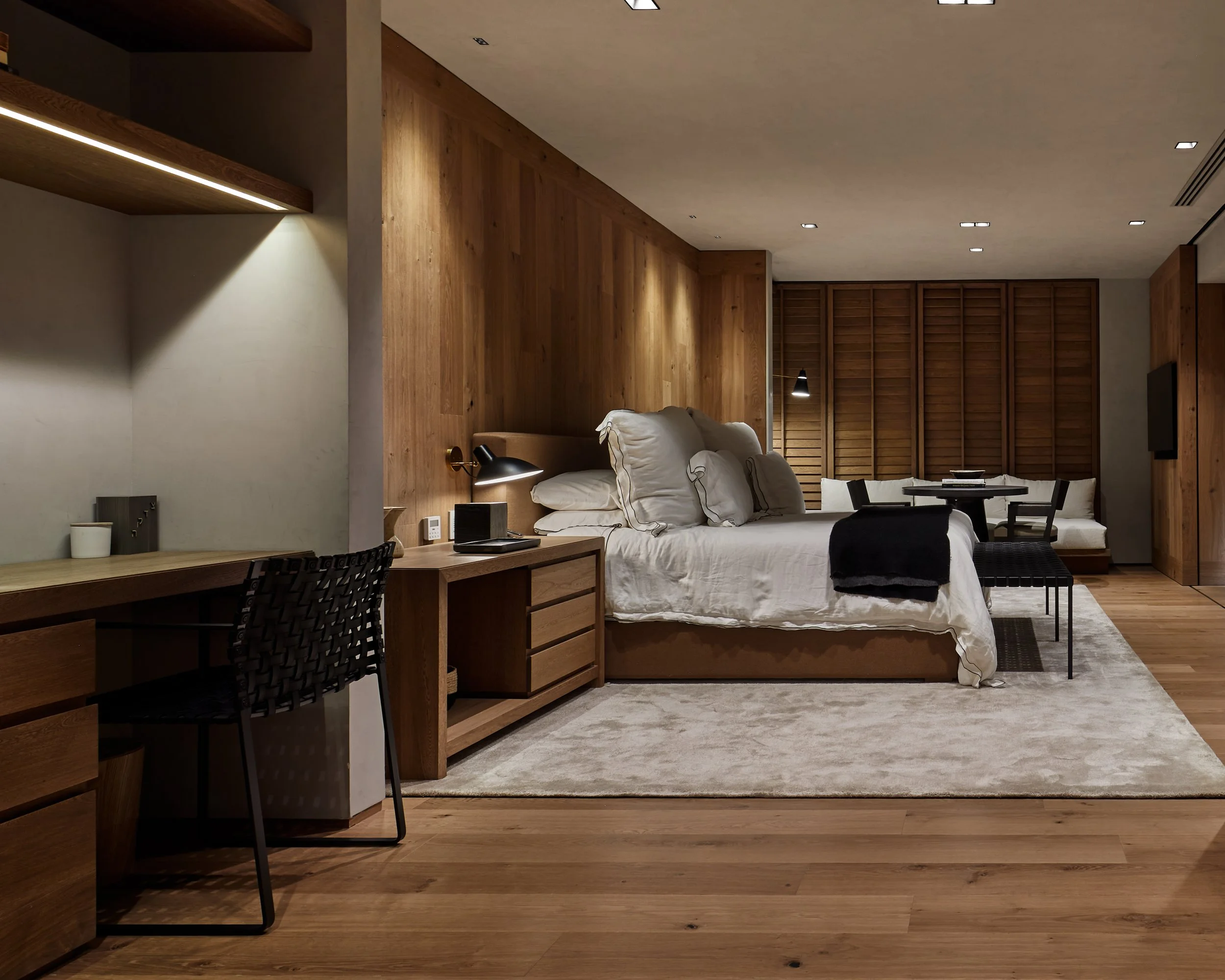 Habitación moderna con paredes y suelo de madera, cama con diversas almohadas blancas, área de trabajo con escritorio y silla de diseño, y área de comedor con mesa y sillas blancas, iluminación suave y muebles de madera.
