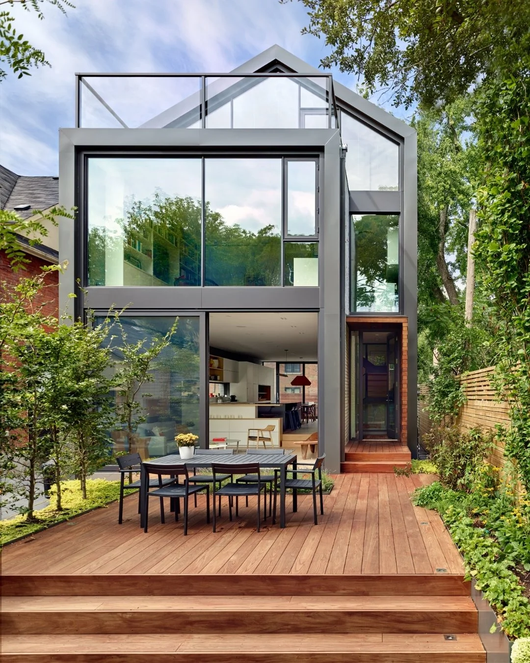Casa moderna de dos pisos con paredes de vidrio grande, jardín con plantas y árboles, y área de comedor exterior con mesa y sillas de madera en un entorno verde