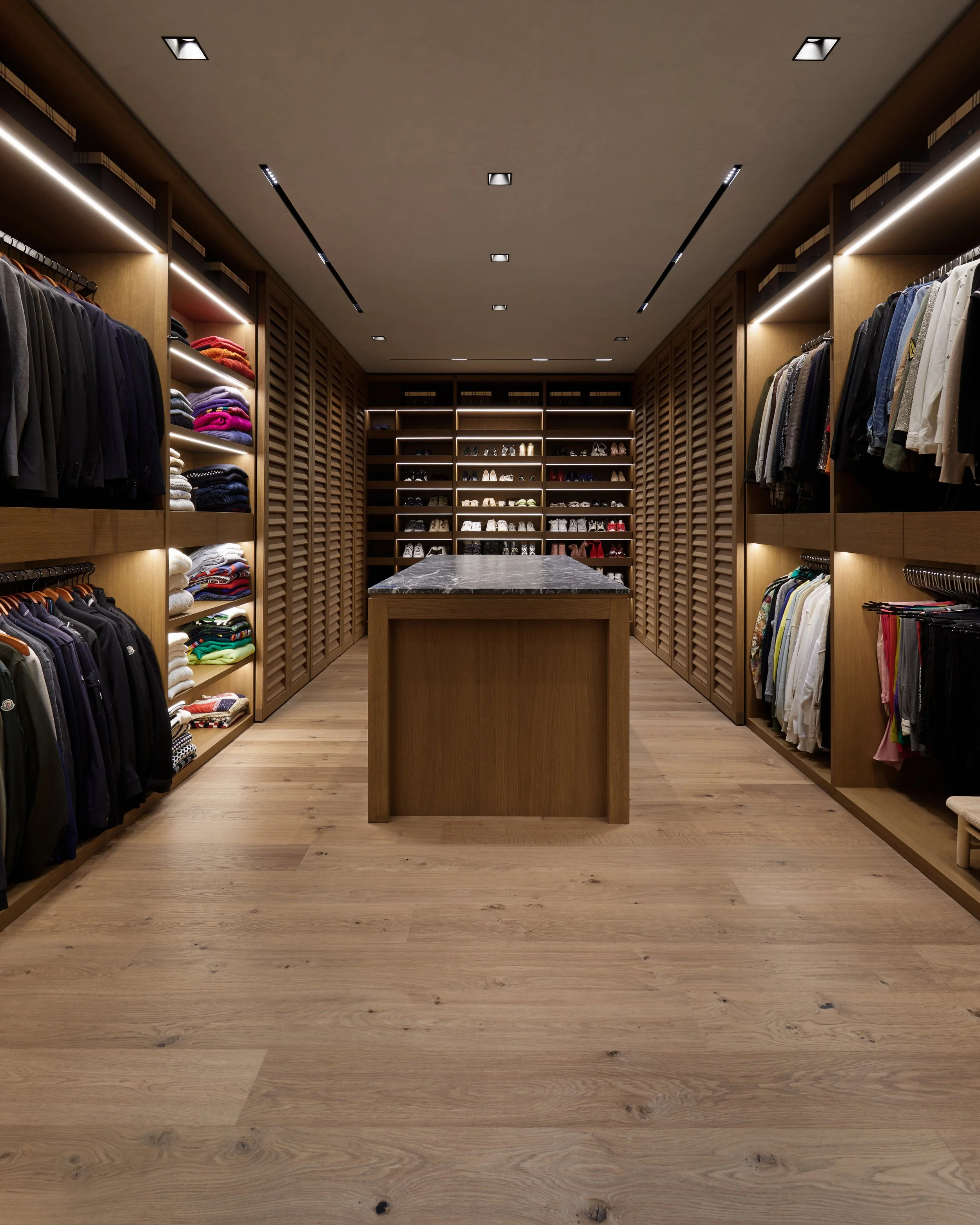 Vestidor bien organizado con ropa y calzado en estantes de madera y una mesa de mármol en el centro.