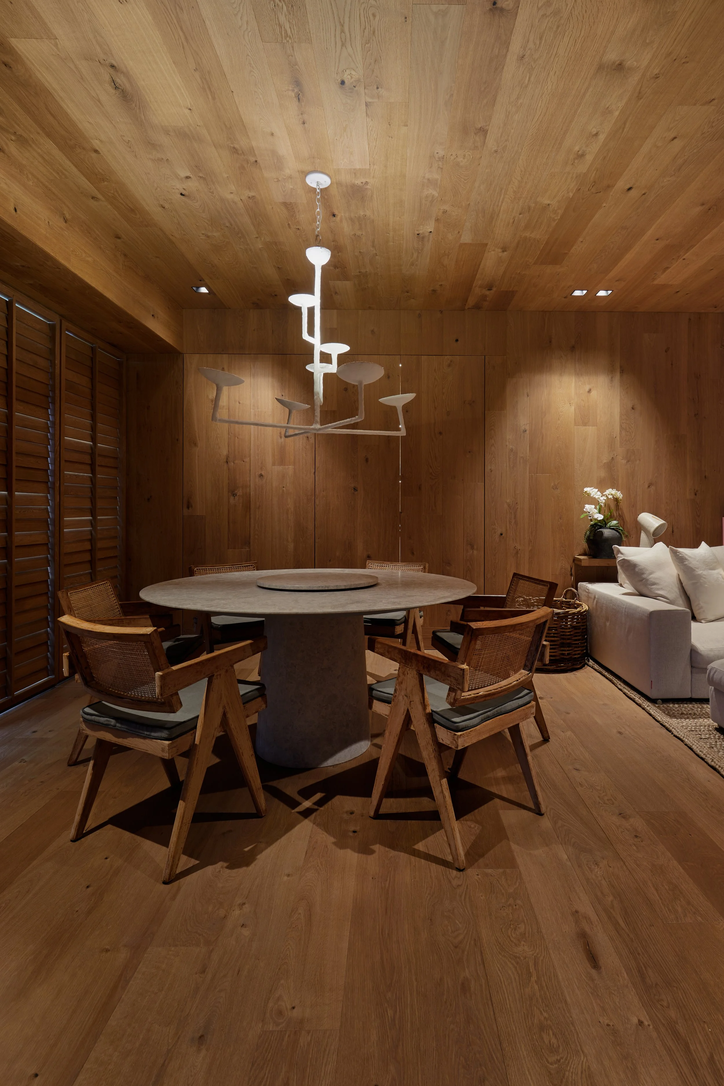 Sala de comedor con paredes y techo de madera, muebles de madera, un comedor redondo con sillas, una lámpara moderna y un sofá blanco con cojines y una lámpara de escritorio, plantas y una alfombra.