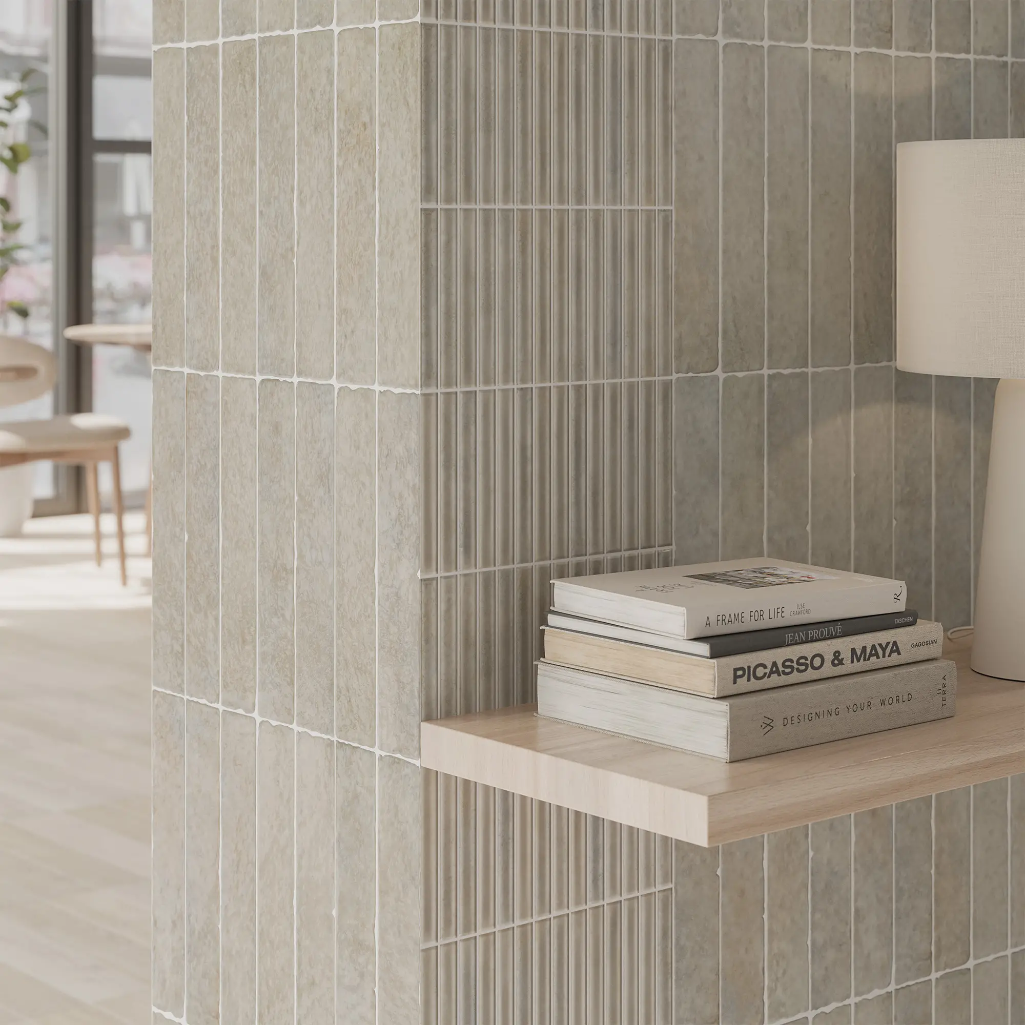 Un estante de madera con libros en una pared de azulejos gris claro. Se ve una lámpara blanca y una ventana con vista a una sala con mesas y sillas de madera.