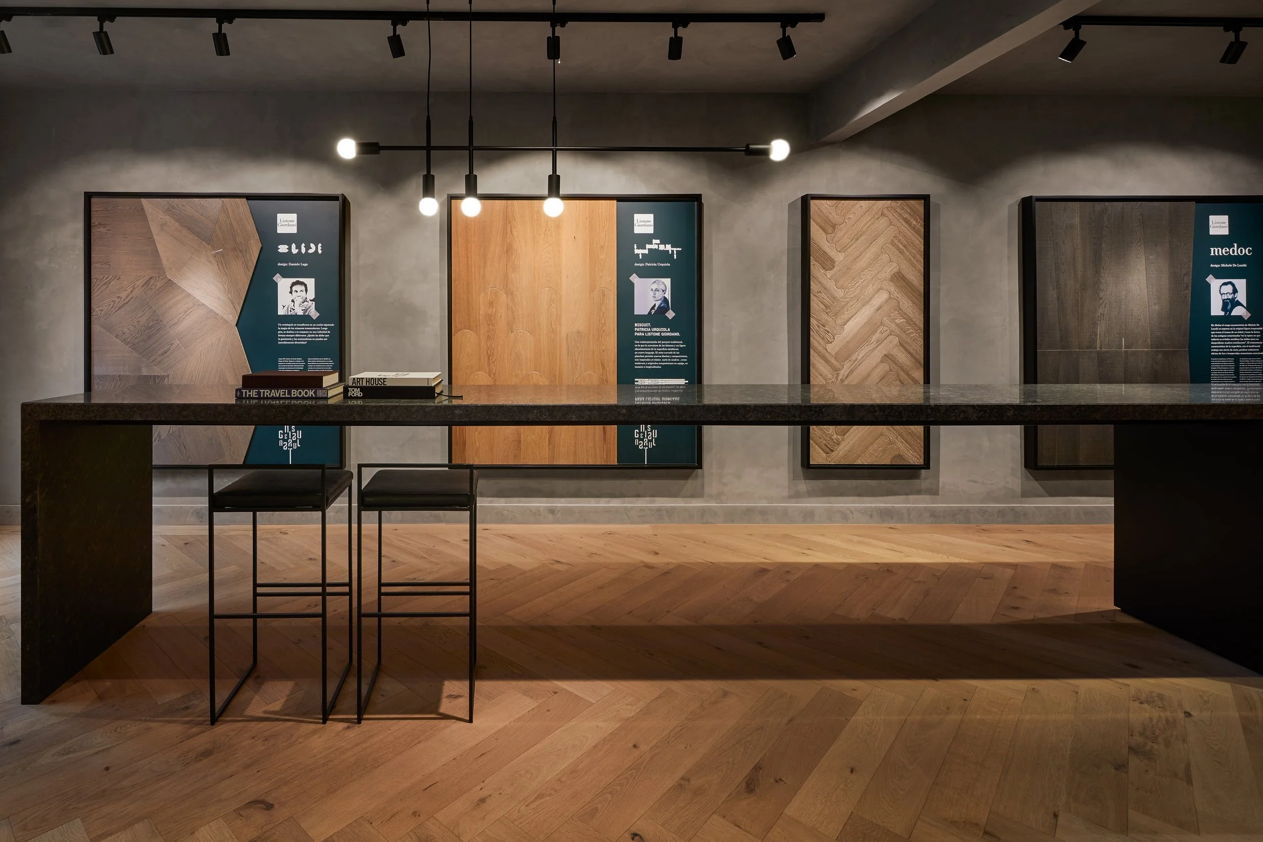 Exposición en una galería con varias láminas decorativas de madera enmarcadas colgadas en la pared. En el centro, una mesa negra con algunos libros y dos sillas altas negras. La iluminación es moderna, con focos y lámparas que iluminan las láminas.