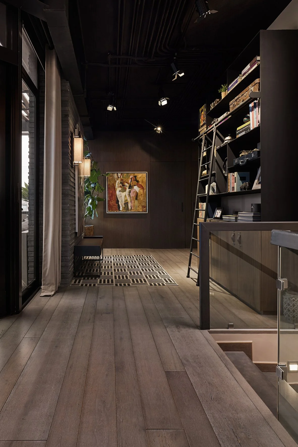 Interior moderno con piso de madera clara, paredes oscuras y estantes llenos de libros, una escalera para acceder a la parte superior de la librería, una pintura en la pared y plantas decorativas.
