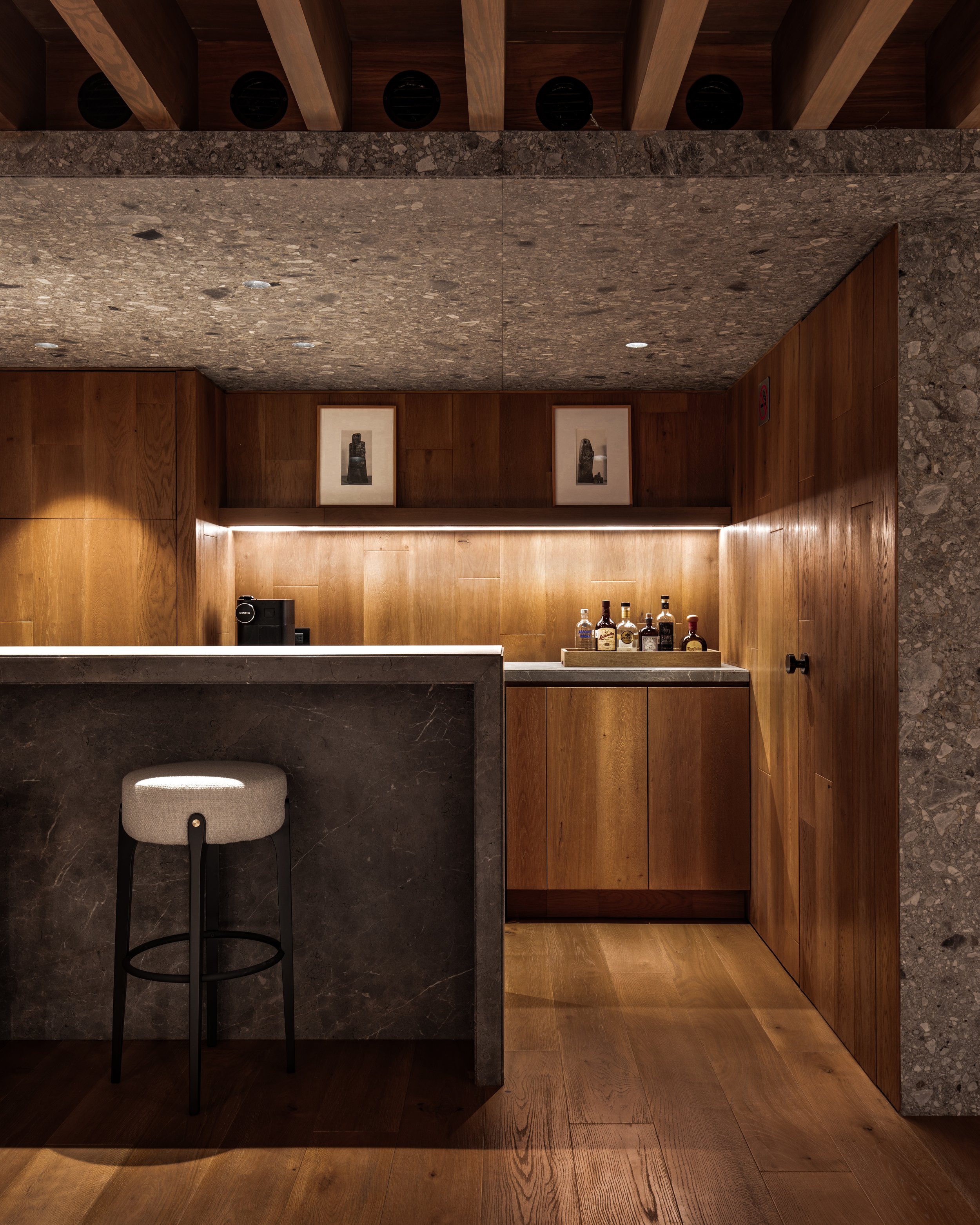 Bar de madera con botellas de licor y una barra de piedra gris, con taburete de asiento beige y respaldo gris, iluminación cálida, cuadros en la pared de madera y techos de madera y piedra.