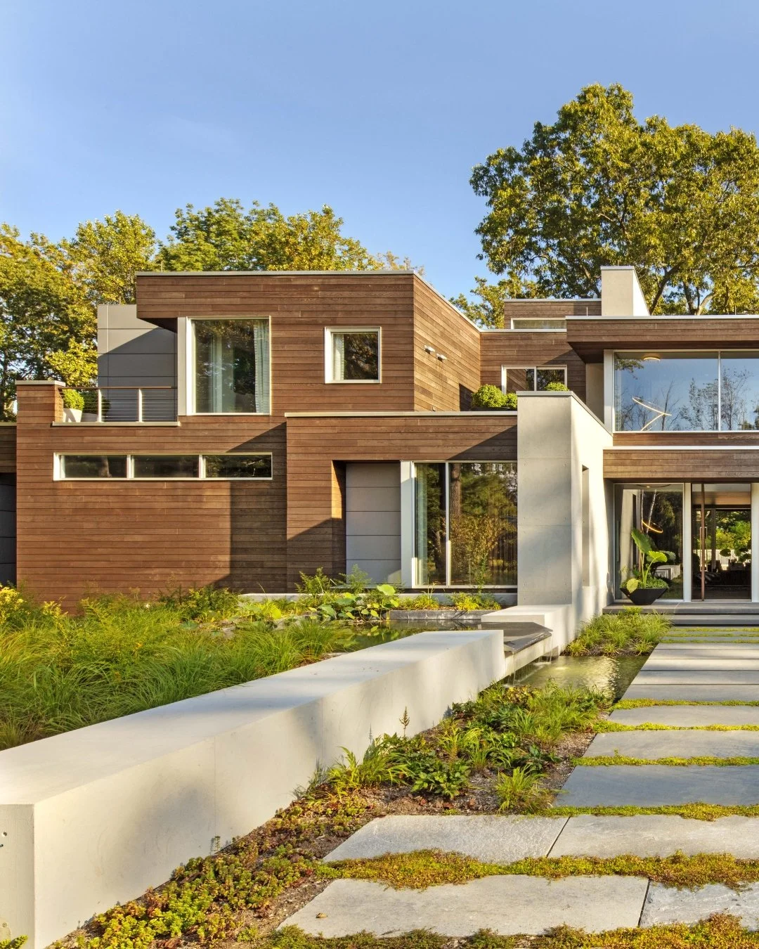 Casa moderna con fachada de madera y grandes ventanas, rodeada de vegetación y un camino de piedra.