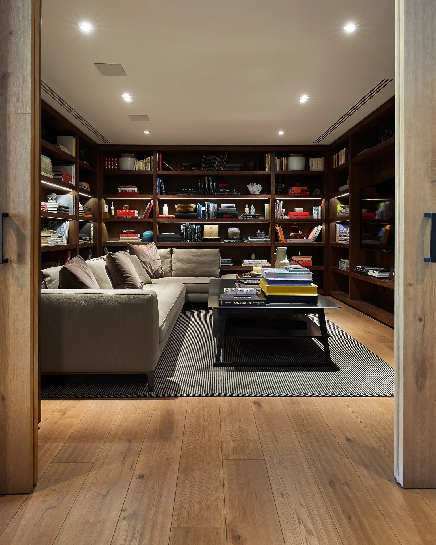 Sala de estar con librerías de madera llenas de libros, un sofá beige con cojines, una mesa de centro con libros apilados y una alfombra a rayas sobre un piso de madera.