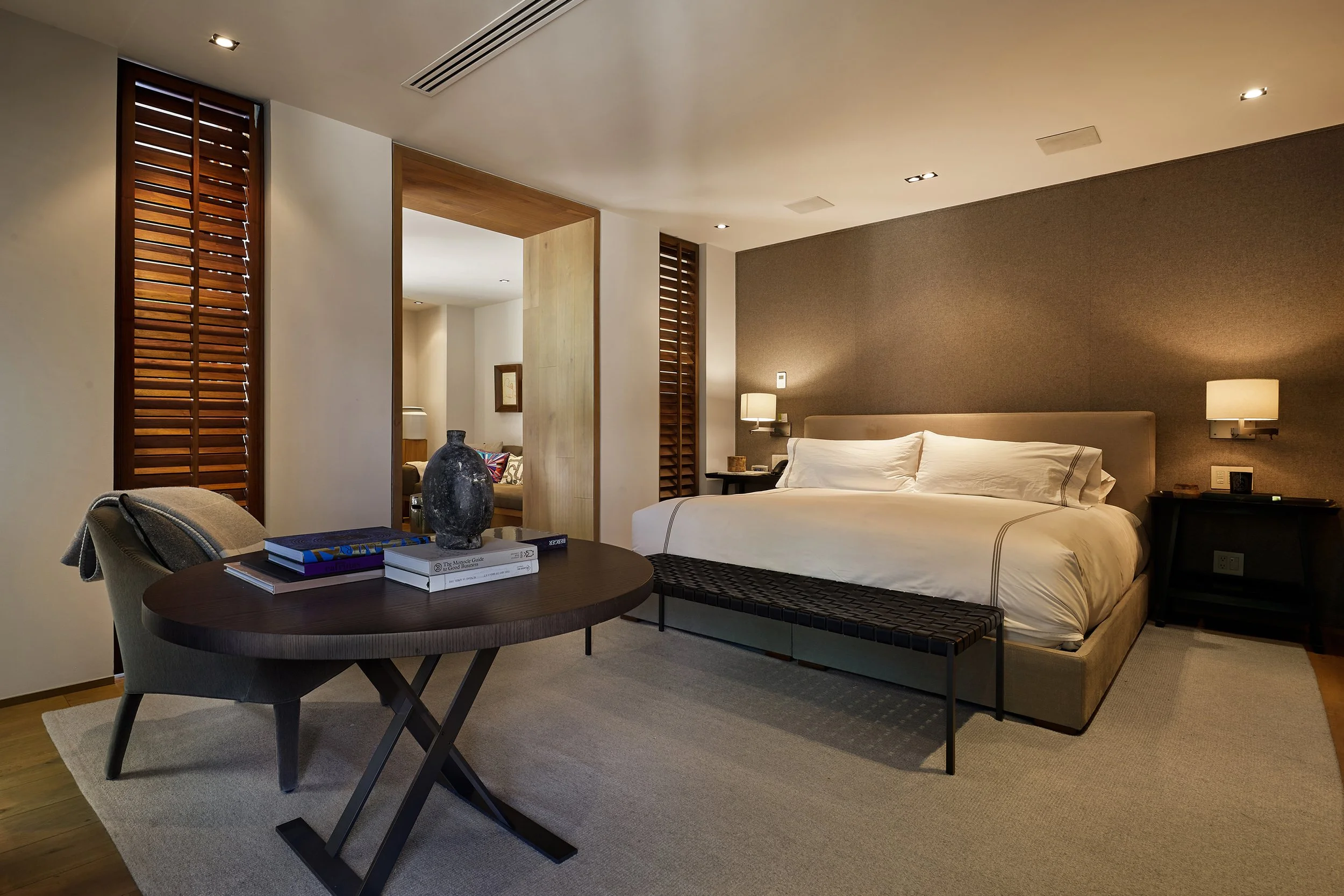 Habitación de hotel con cama king-sized, sillas, mesa con libros y decoraciones, paredes de colores cálidos, iluminación suave y diseño moderno.