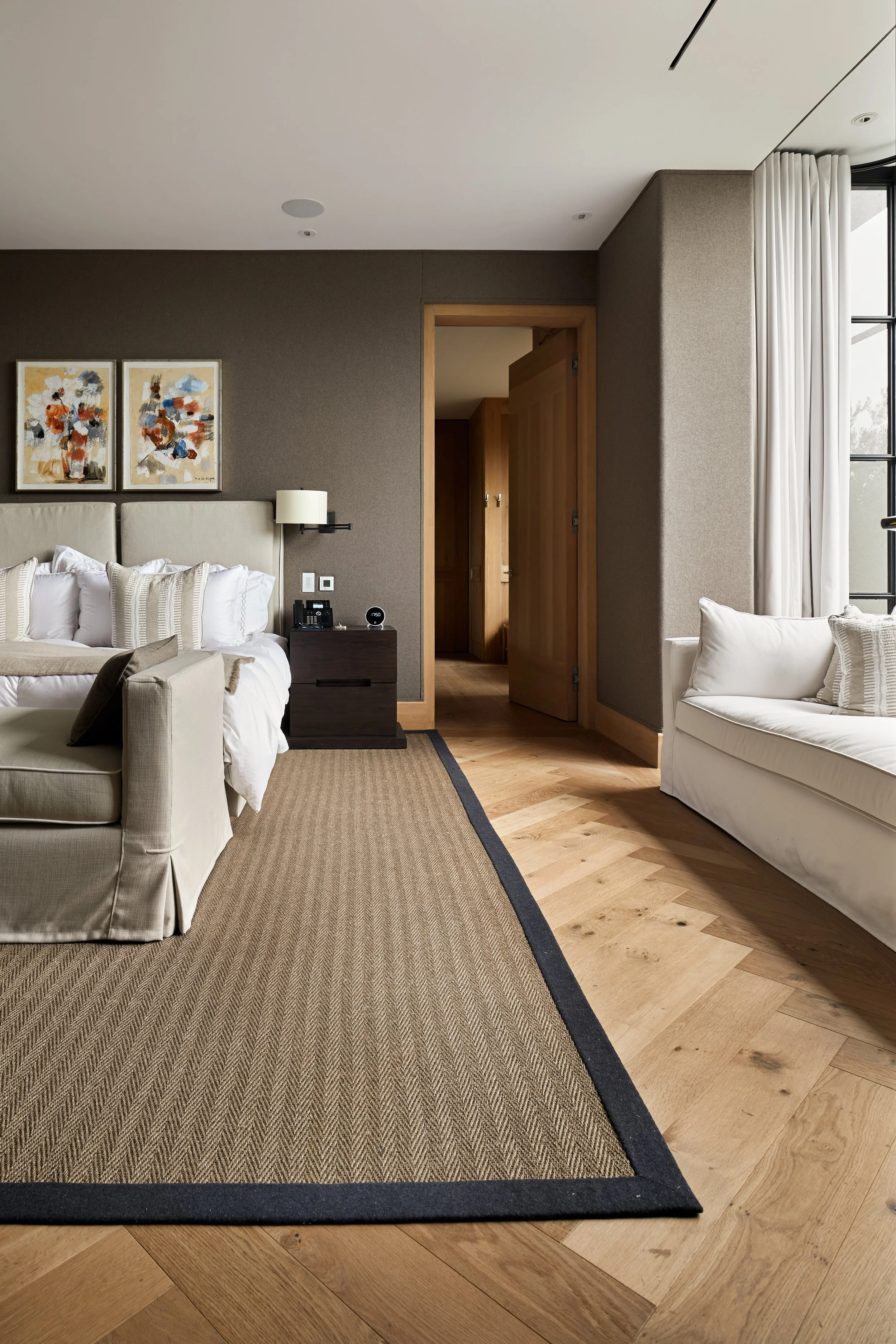 Habitación de hotel con sillón beige, sofá blanco, alfombra beige, paredes de color marrón, instalación moderna, ventana grande con cortinas blancas, arte abstracto en la pared.