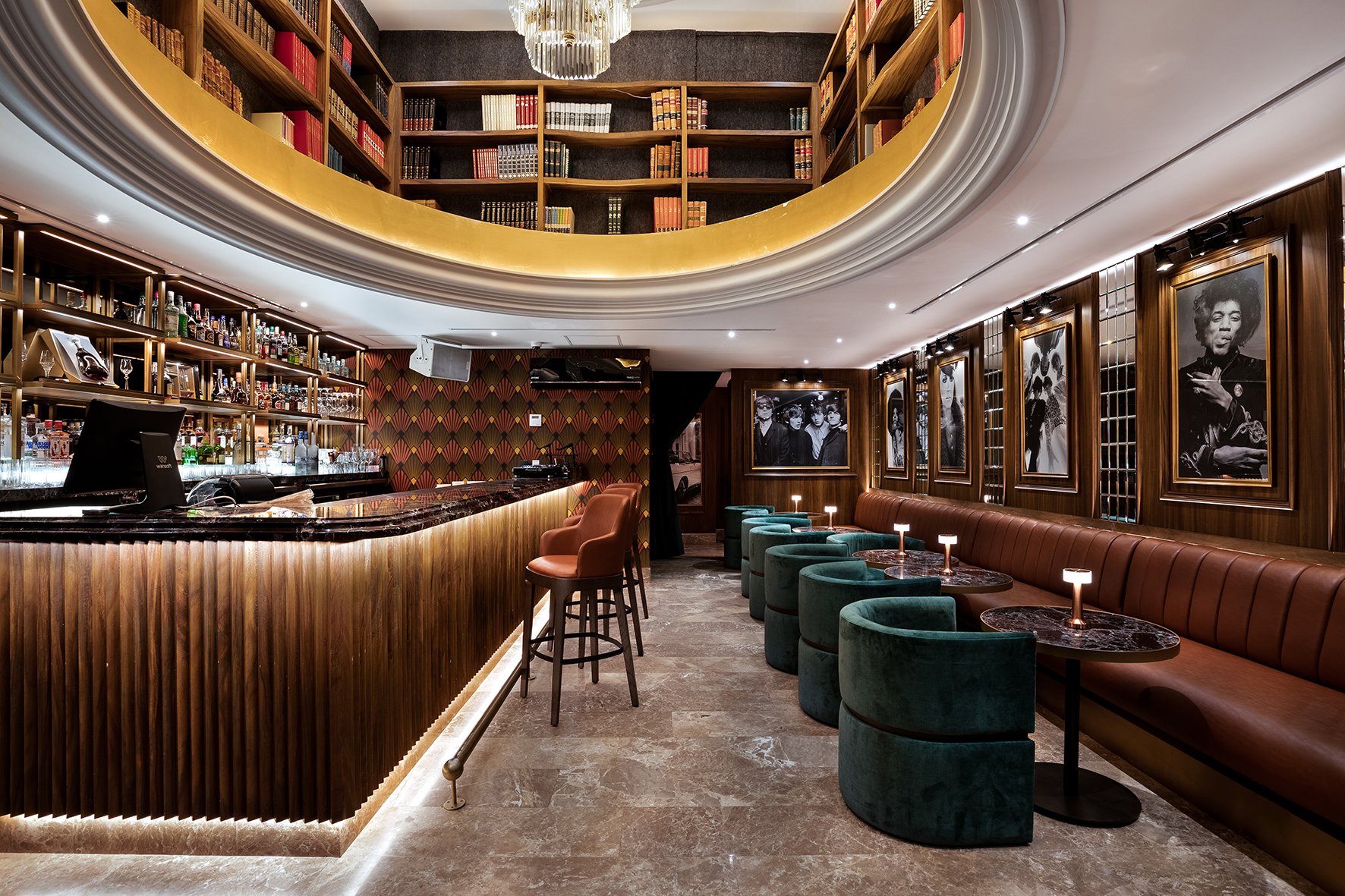 Interior de un bar elegante con barra de madera, sillones de respaldo naranja y sillas verdes, paredes de madera con cuadros en blanco y negro, estanterías con libros en una terraza circular en la parte superior y una decoración sofisticada.