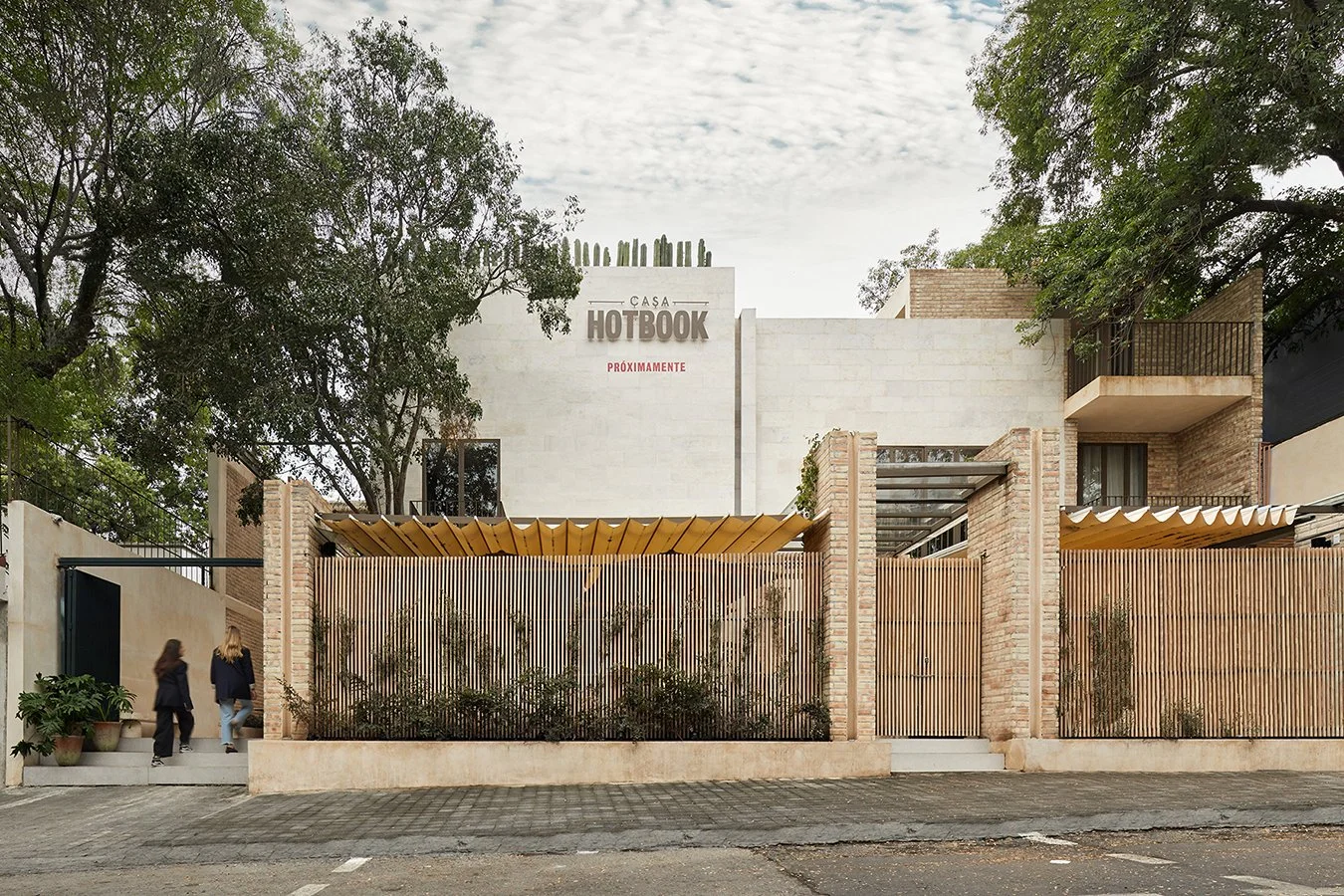 fachada moderna de edificio con muro de bloques de ladrillo, valla de madera, árboles y dos personas caminando