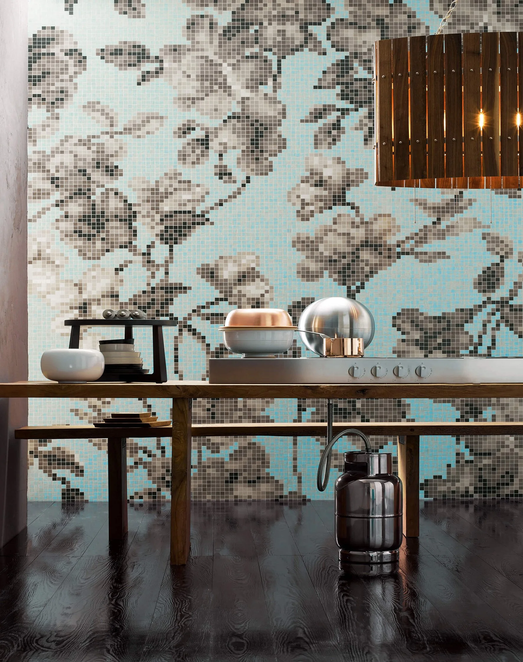 Decoración de cocina moderna con mosaico de fondo con patrón de hojas en tonos grises y azules, una mesa de madera con utensilios metálicos y cerámicos, y una lámpara de madera colgante.