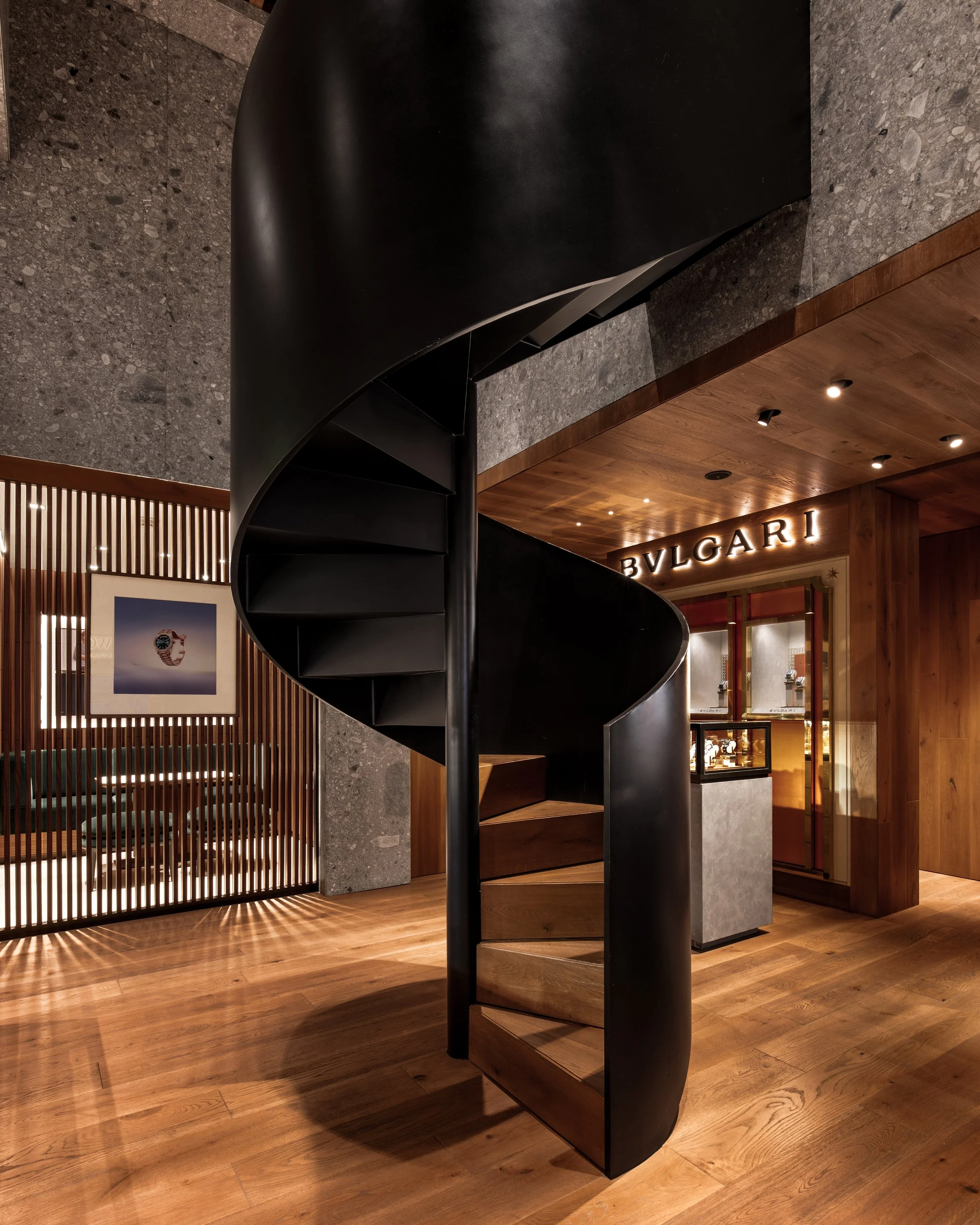 Escalera en espiral negra en un interior de diseño moderno, con piso de madera y paredes de piedra, y una tienda de lujo visible en el fondo.