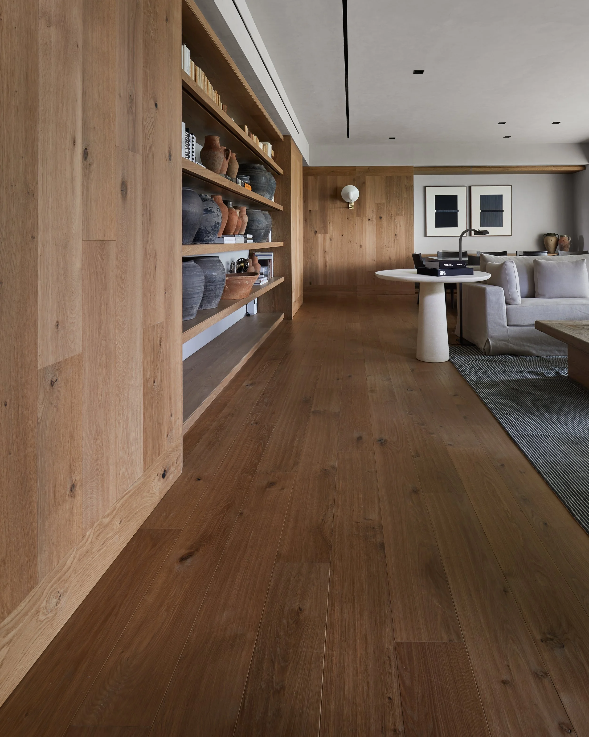 Interior moderno con paredes y suelo de madera, estantería con vasijas y libros, sofá gris, mesa redonda con lámpara, decoraciones en la pared.