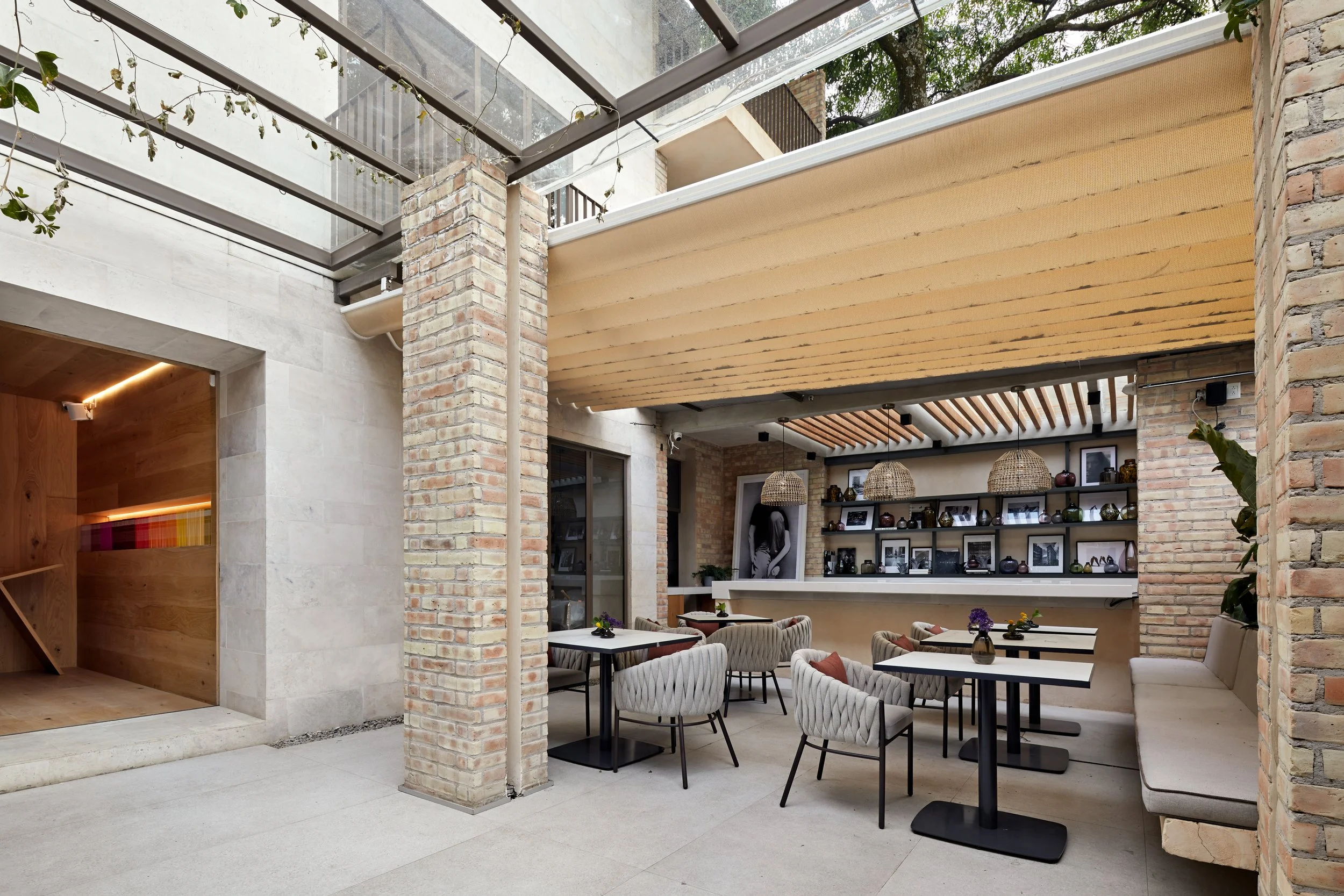 Interior de un restaurante moderno con paredes de ladrillo expuesto, mesas con sillas acolchadas en tonos neutros, y decoraciones en estanterías. Hay techos de madera y lámparas colgantes de mimbre.