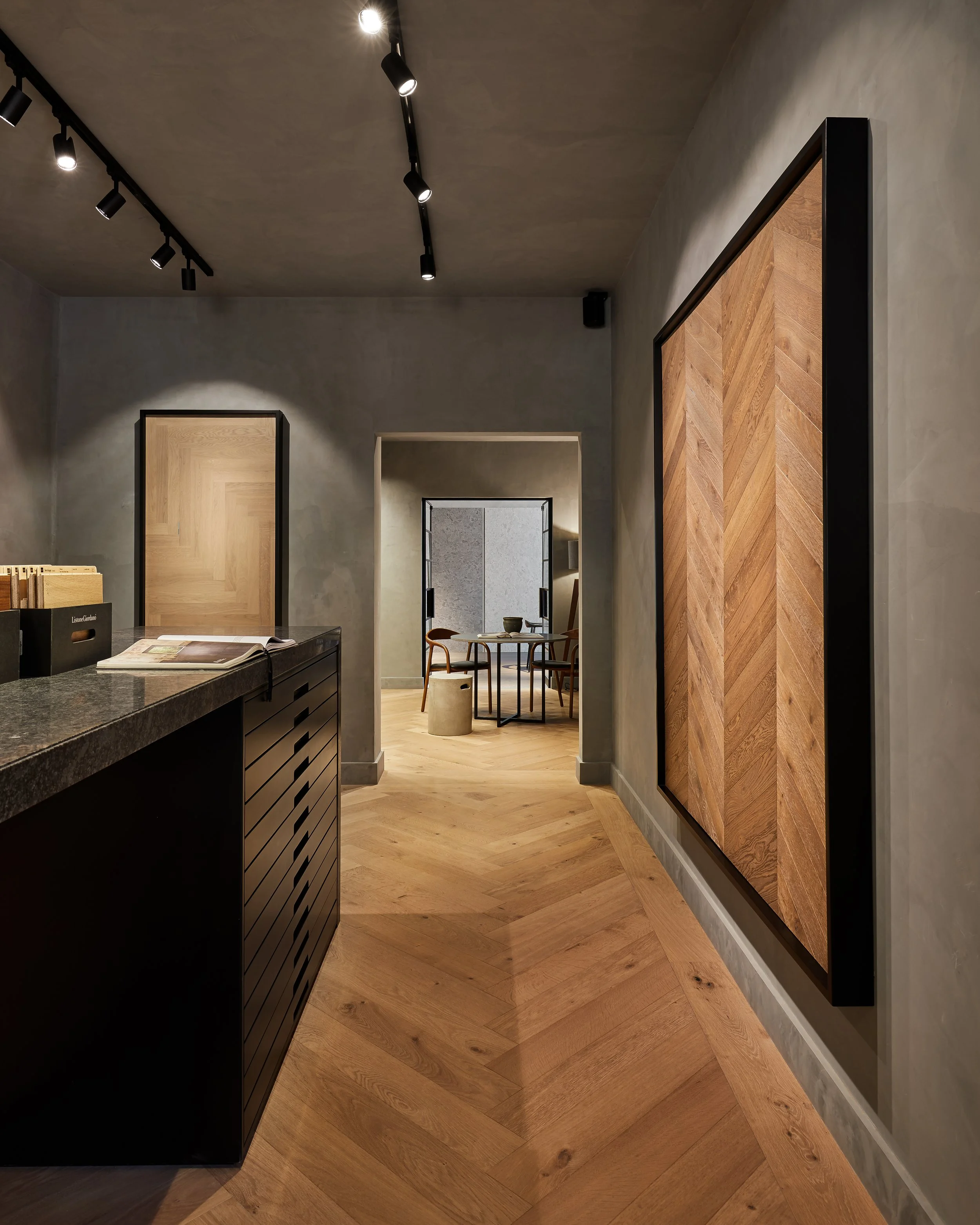 Pasillo de un interior moderno con paredes de color gris, pisos de madera en un patrón diagonales y arte en la pared. En el fondo, una sala de reunión con una mesa y sillas de madera.