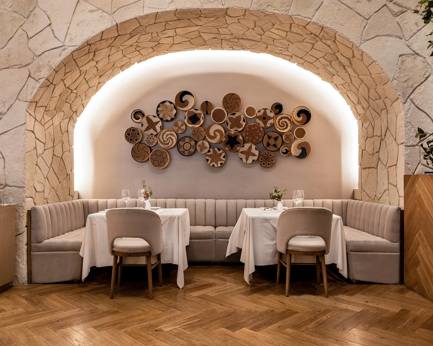 Un espacio de comedor elegante con paredes de piedra y decoración moderna. Hay dos mesas con manteles blancos, una rodeada de sillas y otra de un sofá empotrado. En la pared, arte decorativo hecho de platos con patrones geométricos. La iluminación es