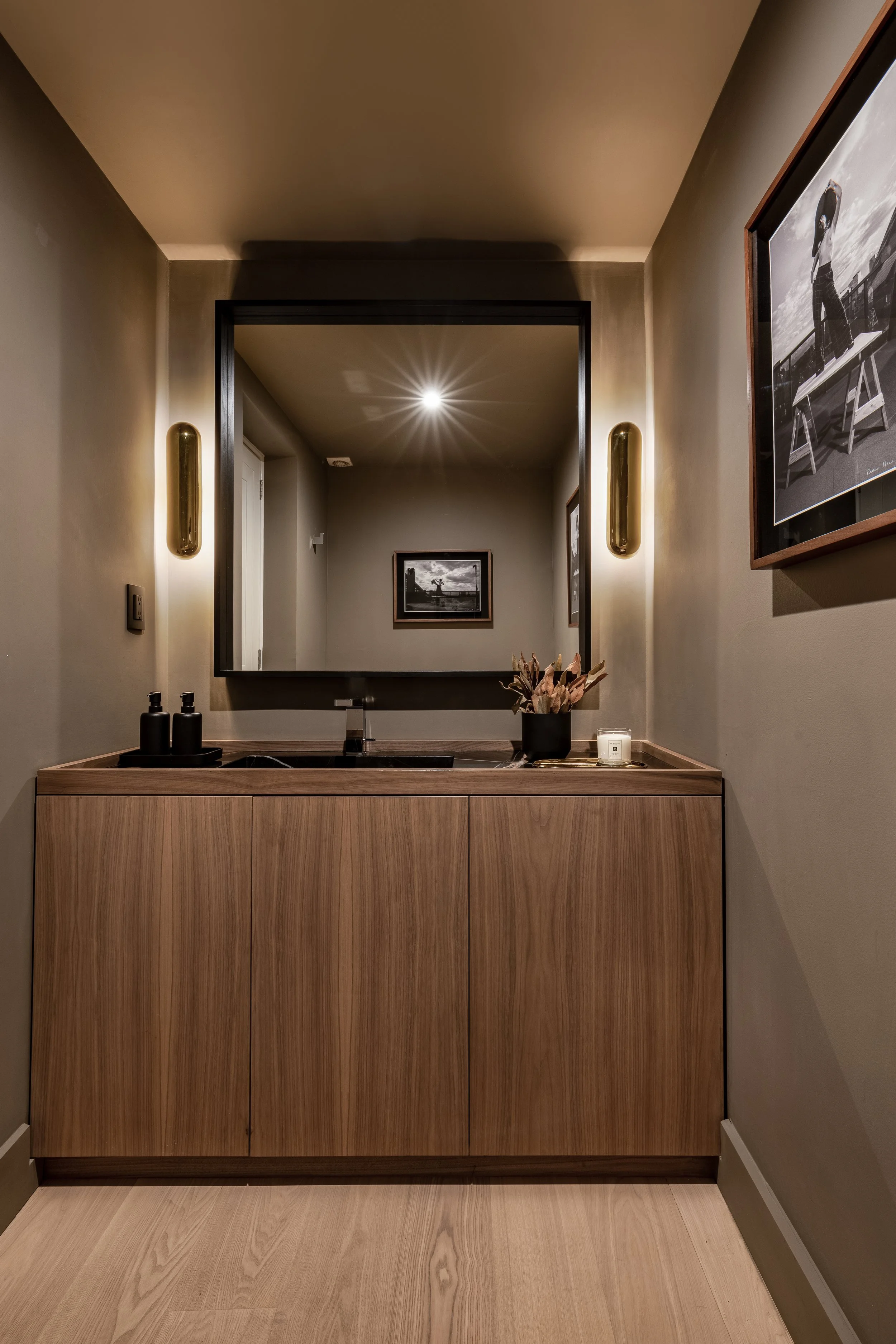 Un lavabo de madera con espejo grande y marcos dorados en una pared gris, con decoraciones como plantas y velas.