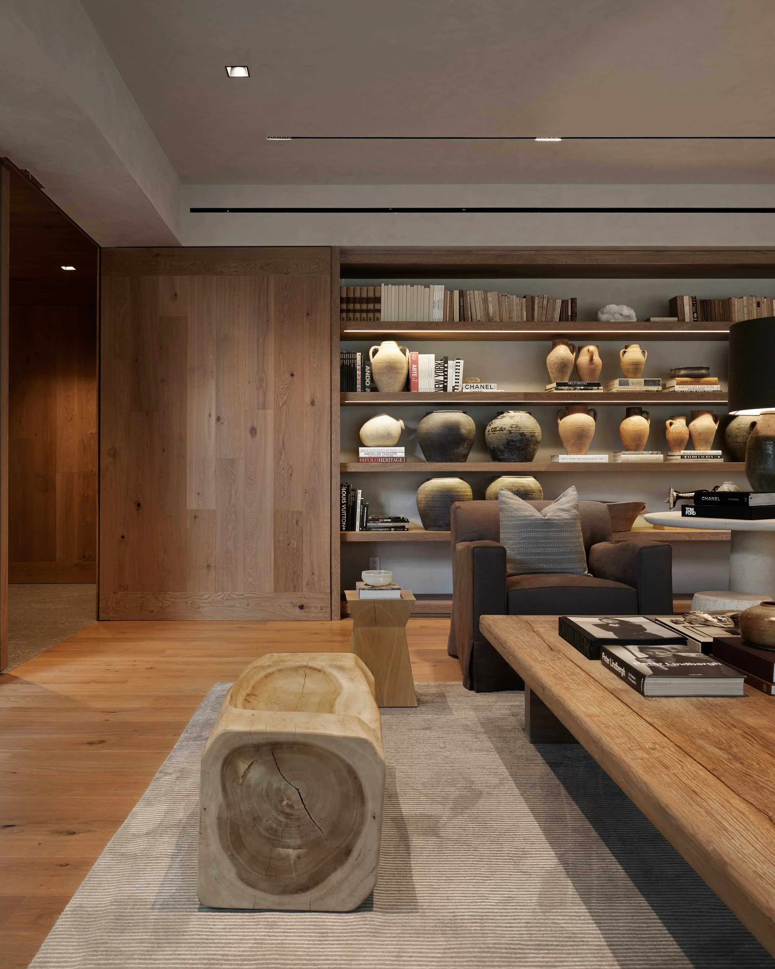 Sala de estar decorada con muebles de madera, una estantería con libros y jarrones de cerámica, sillón marrón con almohada, mesa de madera y alfombra beige.