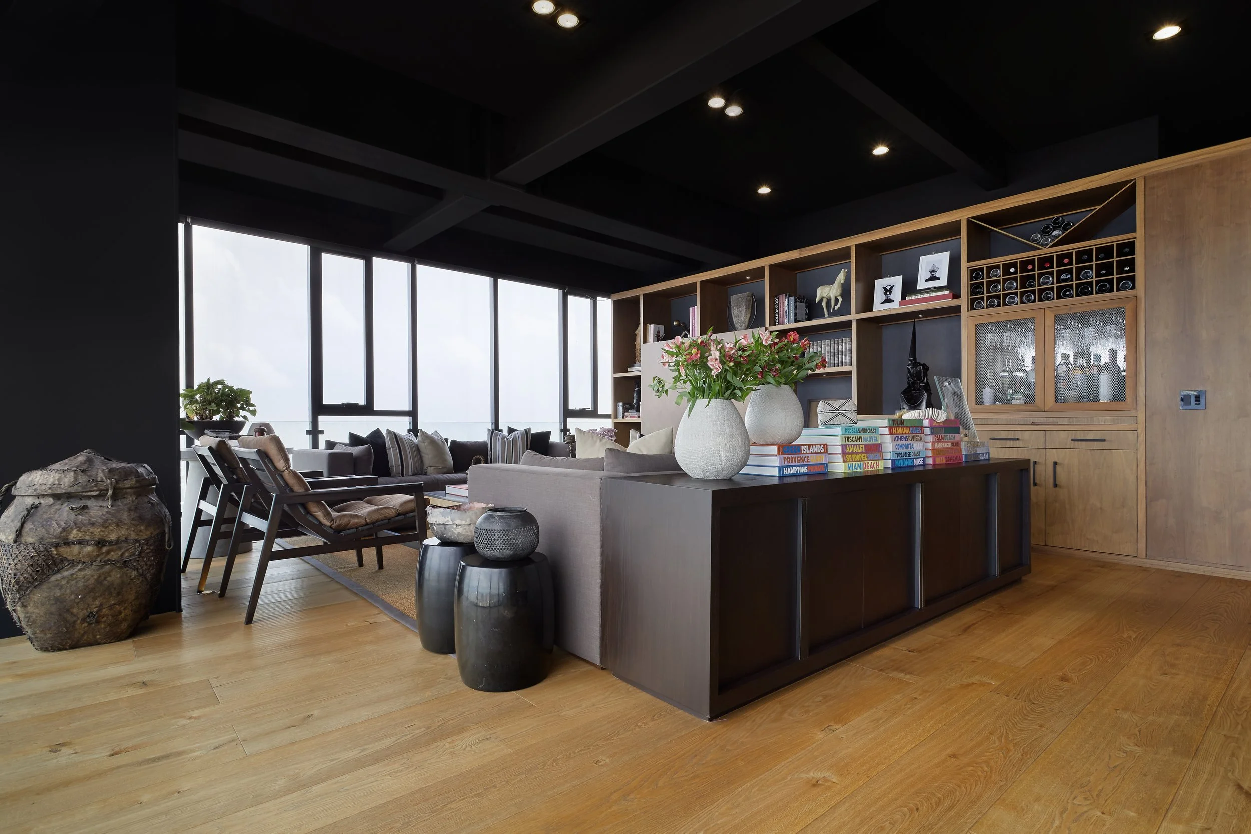 Sala moderna con grandes ventanas, muebles de madera y decoraciones diversas, incluyendo jarrones con flores y estanterías con libros y objetos decorativos.