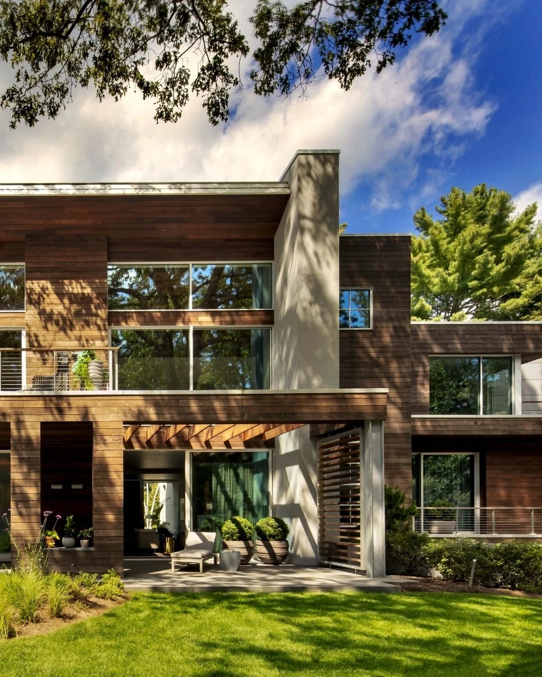 Casa moderna con fachada de madera, ventanas amplias, patio con sillón y plantas, rodeada de césped y árboles, cielo azul con nubes.
