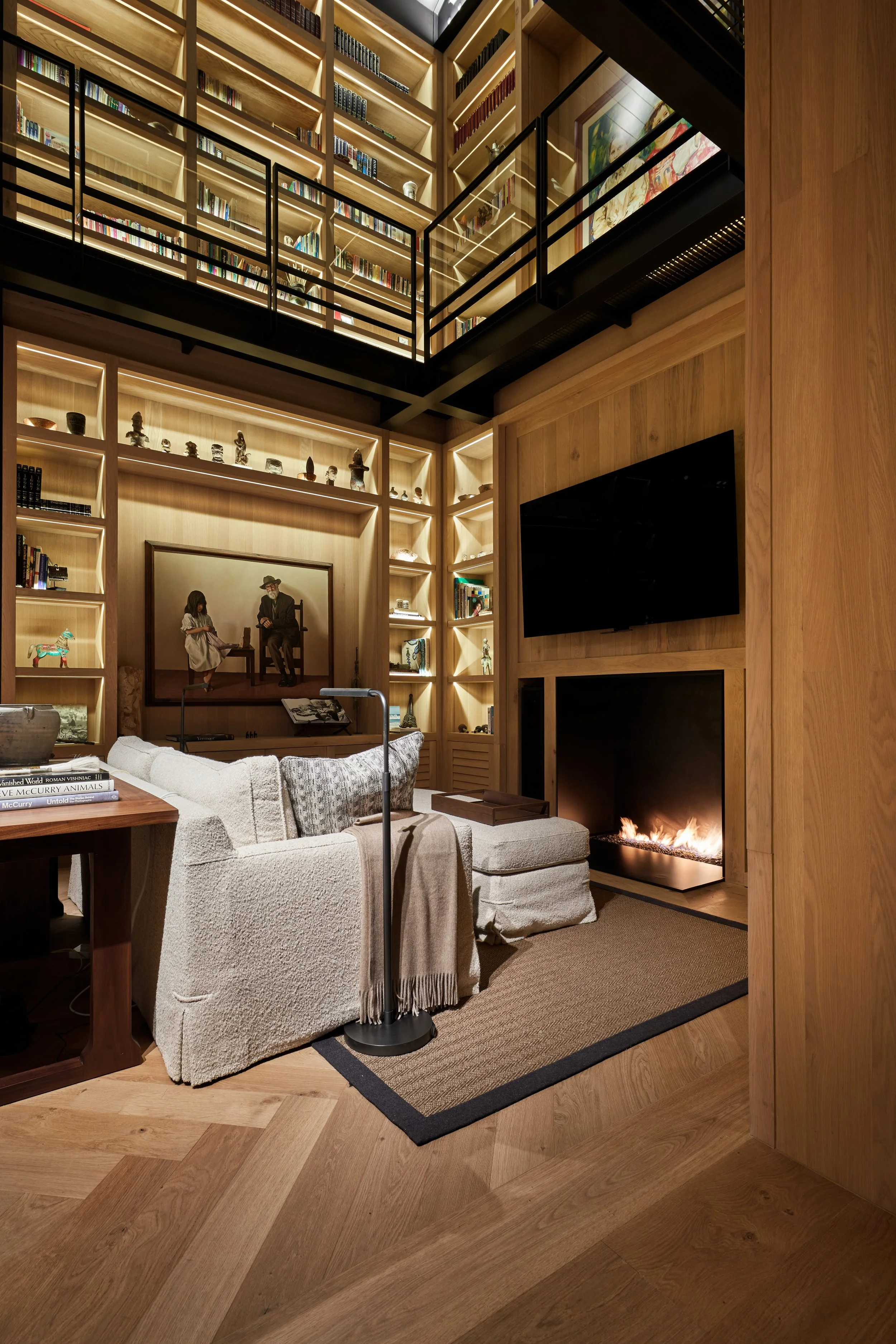 Sala de estar con paredes de madera, estanterías llenas de libros y objetos decorativos, un televisor, una chimenea, un sillón gris y una alfombra.