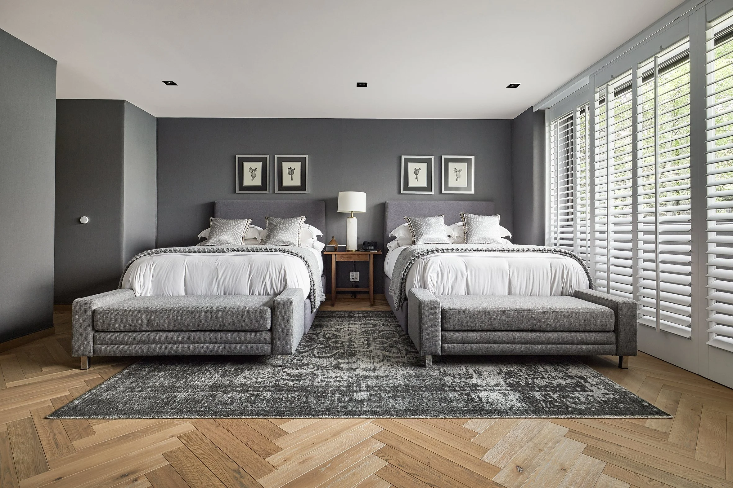 Habitación moderna con dos camas king size, cabeceras grises, decoración en tonos grises y blancos, cortinas blancas con persianas venecianas, alfombra gris y piso de madera.