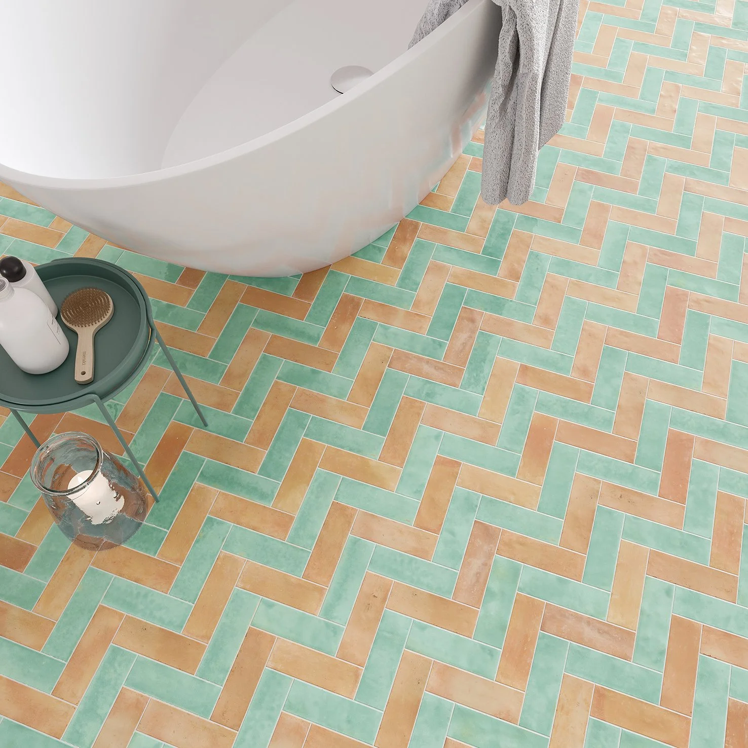 Piso de baldosas en tonos terracota y menta en un baño con bañera blanca, una bandeja con botellas y cepillo de baño, un vaso con agua y una toalla gris colgada.