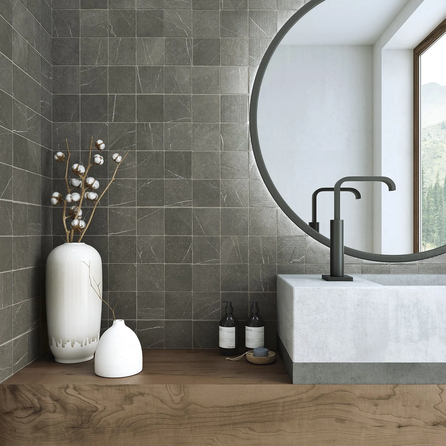 Un lavabo de cemento con una manguera negra, un espejo redondo grande y una ventana que muestra un paisaje natural, en una pared de azulejos gris oscuro. Hay dos botellas de jabón y una pequeña bandeja con un jabón en el mostrador de madera.