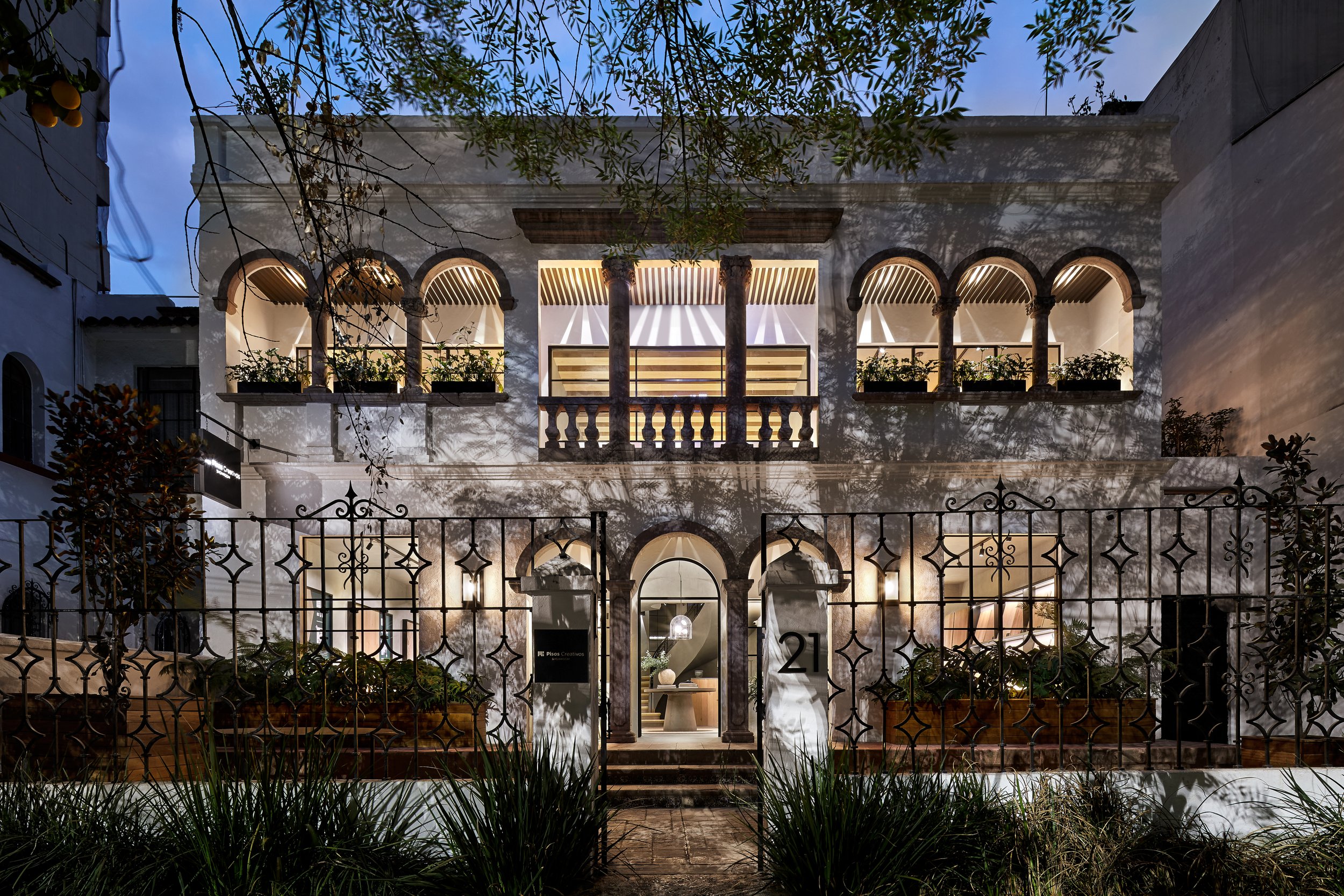 Edificio residencial de arquitectura clásica, con balcones y columnas de piedra, iluminado por luces exteriores por la noche, rodeado de un enrejado de hierro y plantas ornamentales.