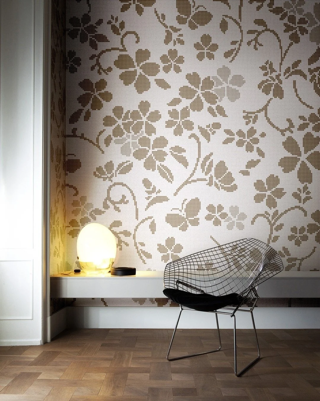 Sala moderna con pared decorativa de mosaico con patrón de flores y hojas en tonos marrón, sillón de estructura de alambre negro con cojín negro, lámpara de mesa en forma de globo que emite luz cálida, en suelo de madera.