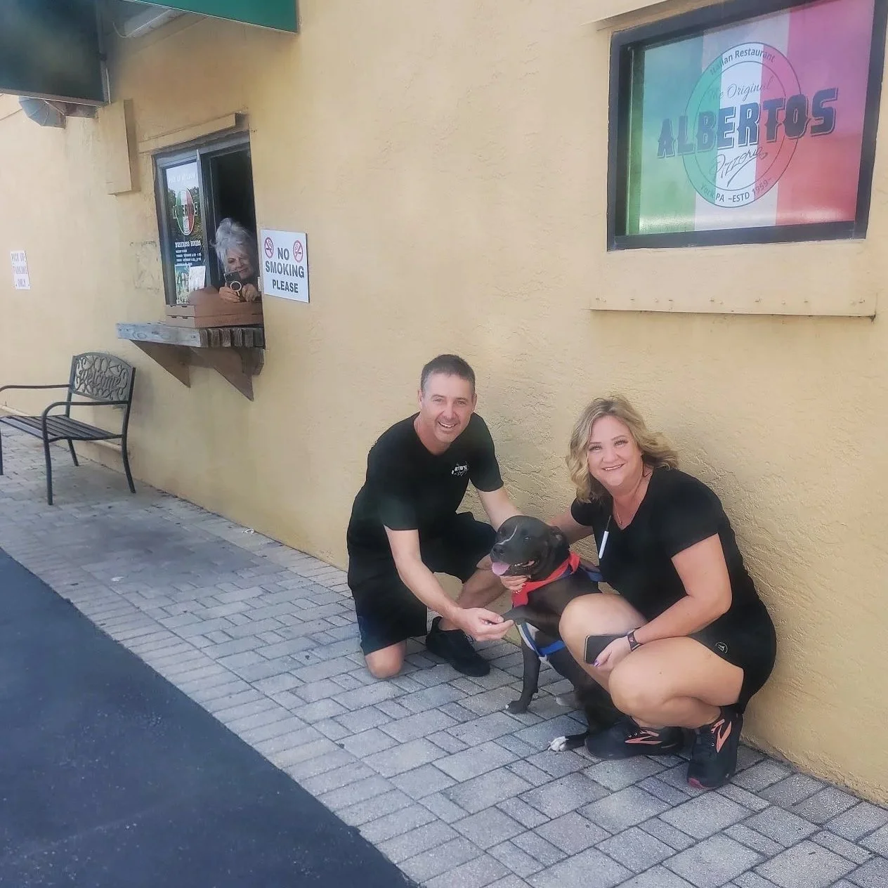 Two smiling people crouching on the sidewalk, holding a black dog with a red bandana in front of a yellow building with a window and a sign that says 'No Smoking Please'.