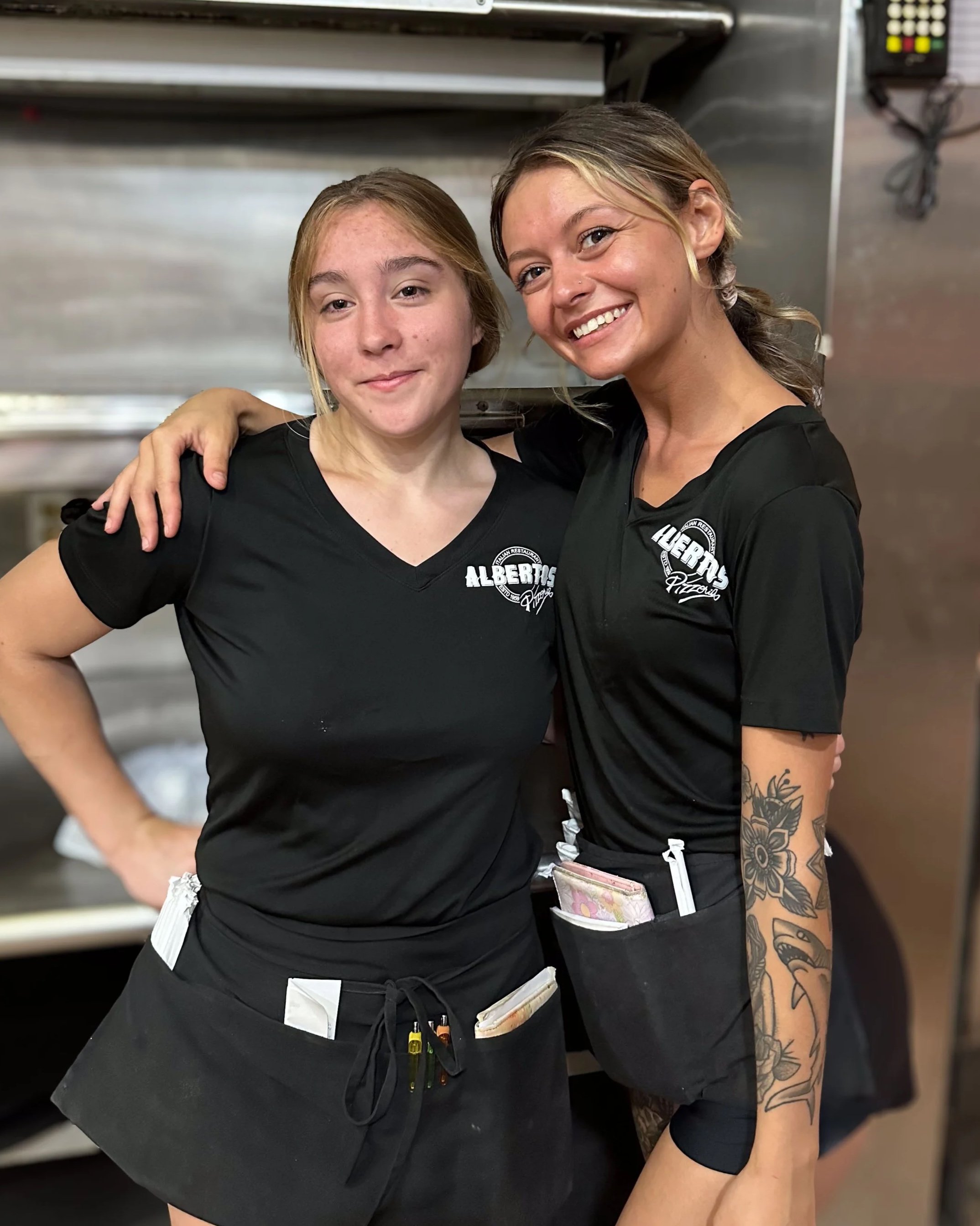 Two women wearing black uniforms with Albertsons logos, smiling and posing inside a commercial kitchen.