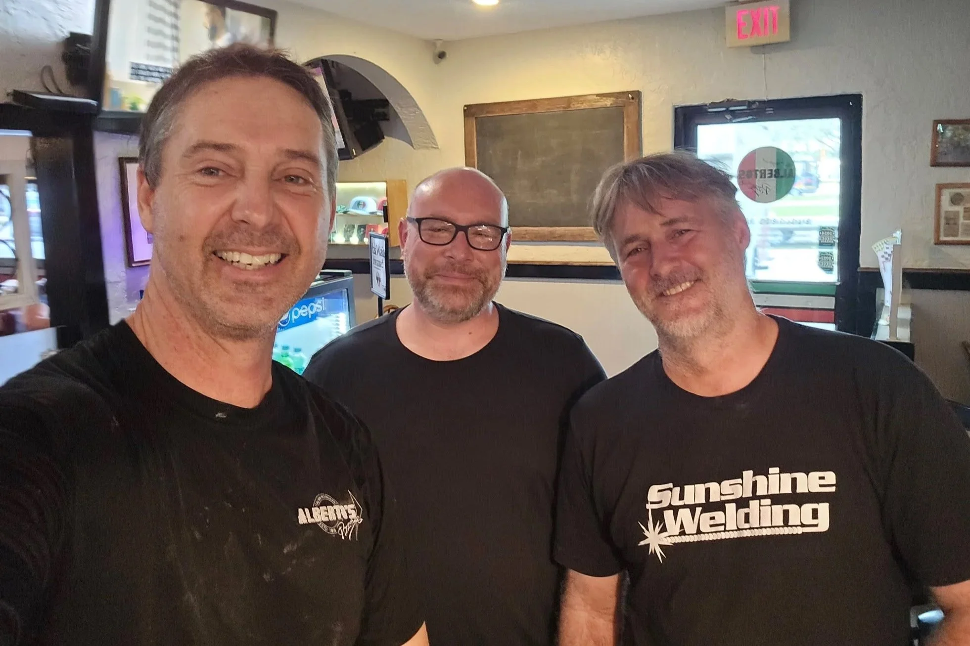 Three men standing inside a restaurant or bar, smiling at the camera. The man on the left has short dark hair and a black shirt with white print. The man in the middle has glasses, a beard, and is wearing a black shirt. The man on the right has gray 