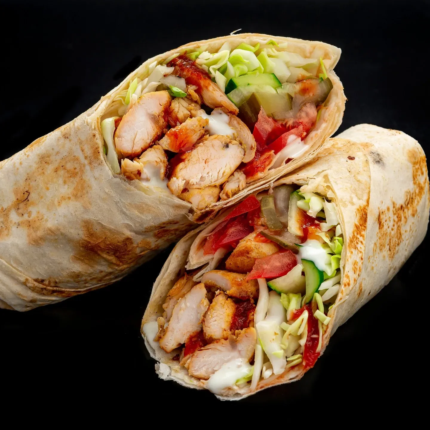 Two burritos sliced in half, filled with grilled chicken, lettuce, tomatoes, cucumbers, and sauce, on a black background.