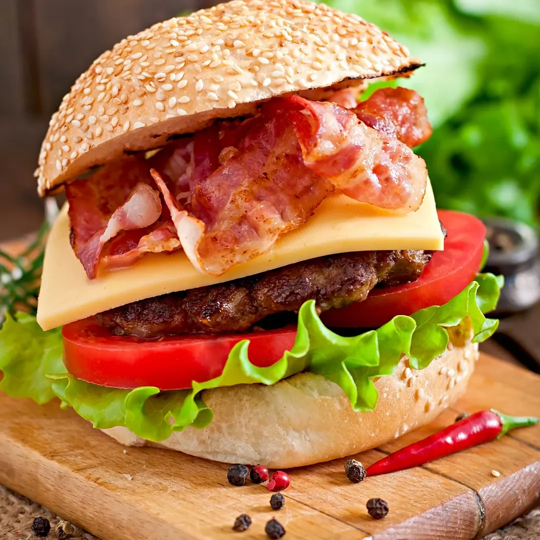 Close-up of a loaded bacon cheeseburger on a wooden cutting board, with lettuce, tomato, cheese, hamburger patty, bacon, and a sesame seed bun, with red chili pepper and peppercorns nearby.
