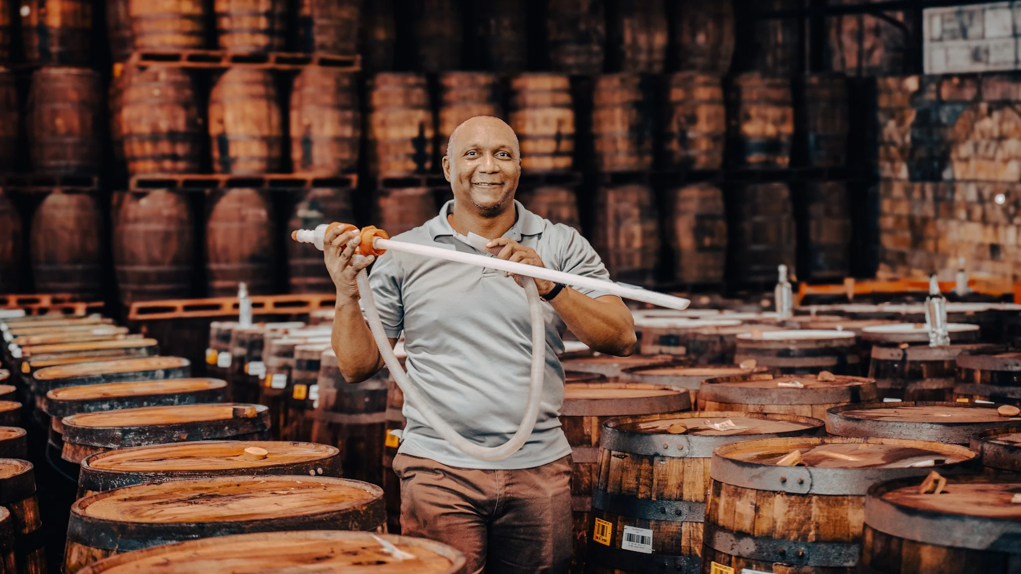 A man smiling while holding a hose in a barrel storage area of a distillery, with rows of wooden barrels in the background.