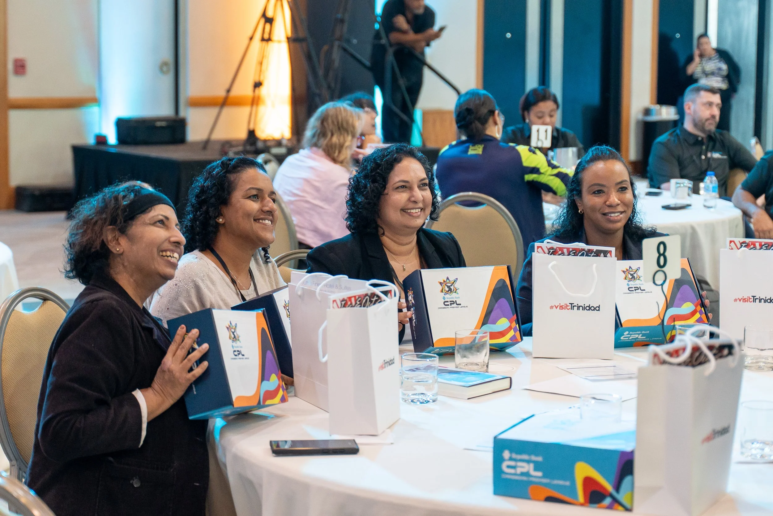 A diverse group of smiling women sitting at a table during a professional event or conference, with branded gift bags in front of them and a few glasses of water on the table.