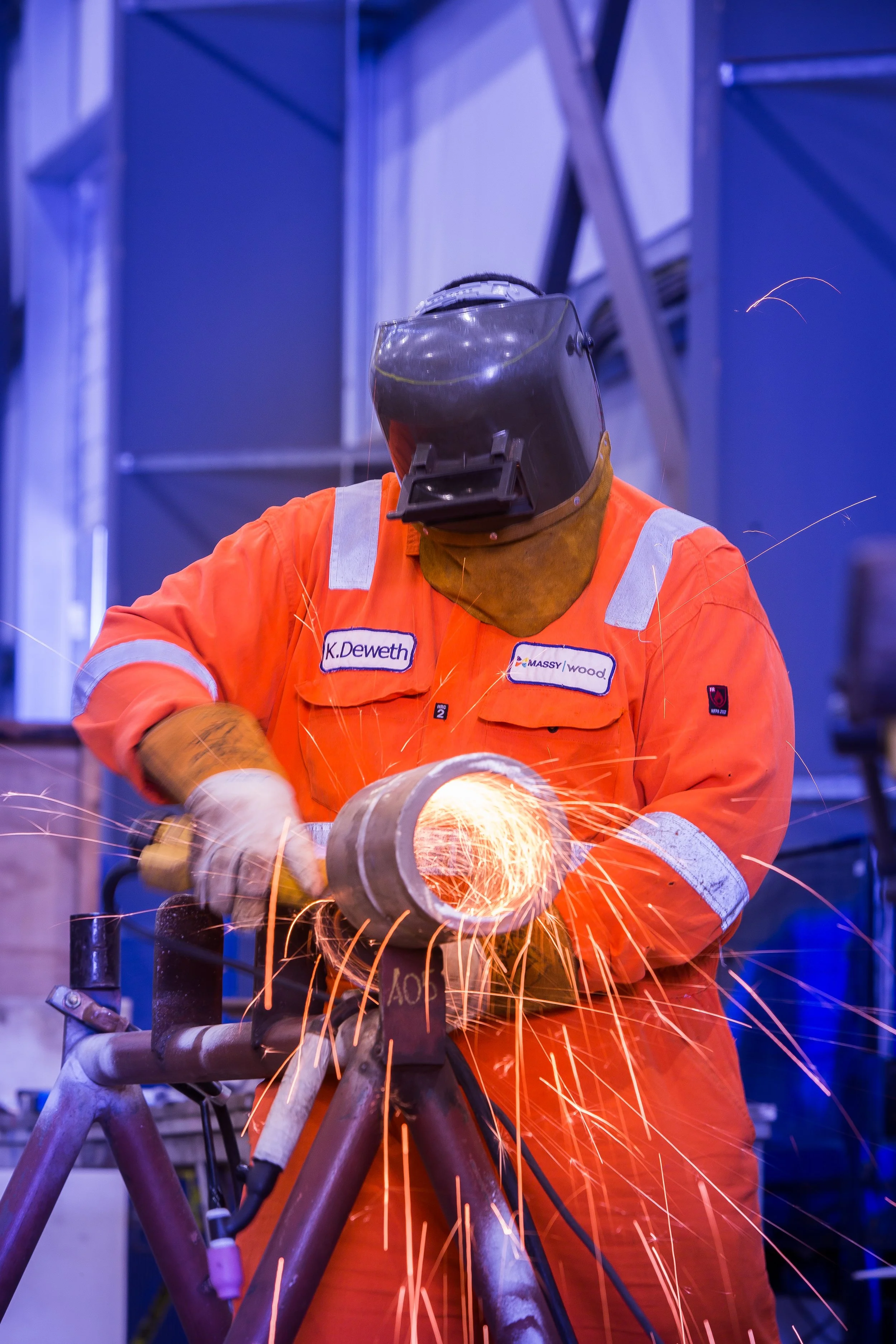 A welder in an orange safety suit and welding helmet is welding a metal structure, producing bright sparks and a small arc of light.