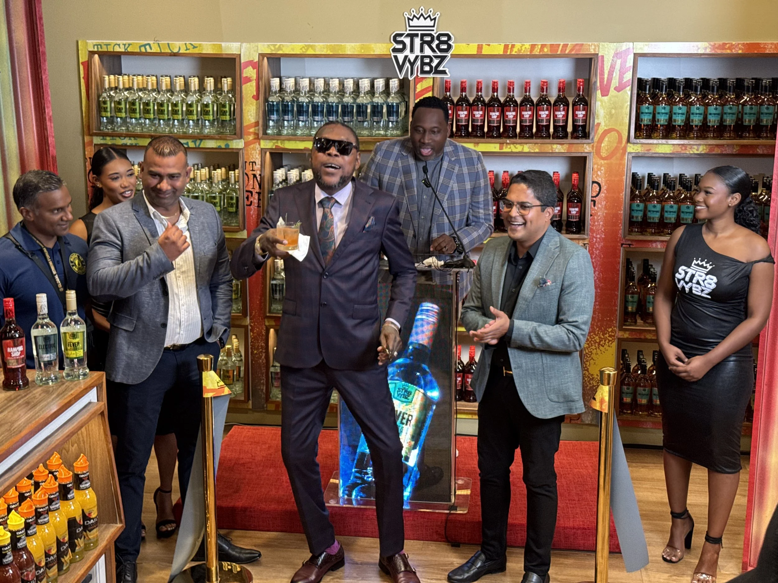 Group of people gathered around a fountain, with some holding drinks, at an event or celebration in a retail store setting with shelves of condiments and sauces.