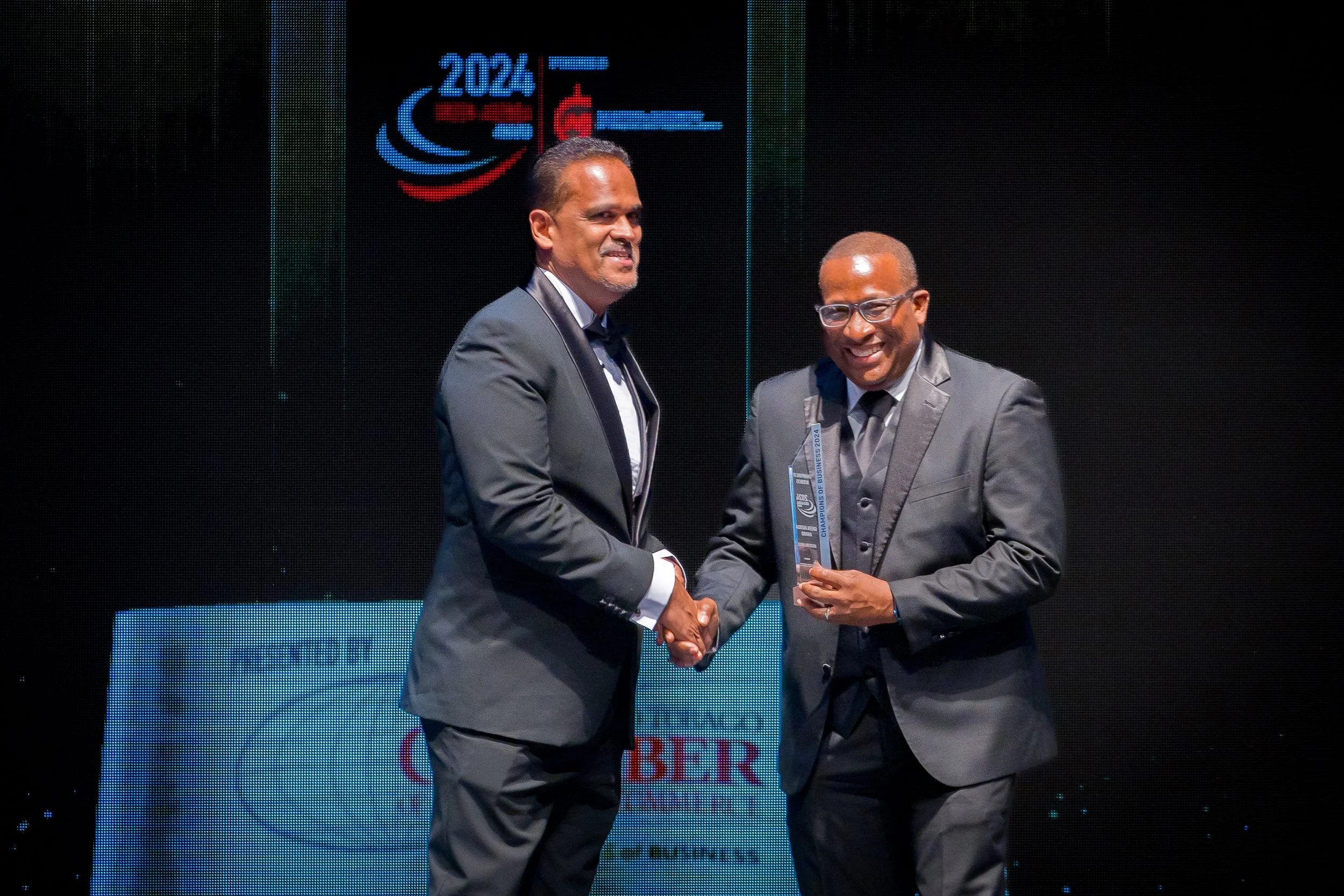 Two men in suits shaking hands at an award ceremony on a stage, with a large digital screen in the background displaying the year 2024 and the word 'Champions of Business 2024' along with logos.