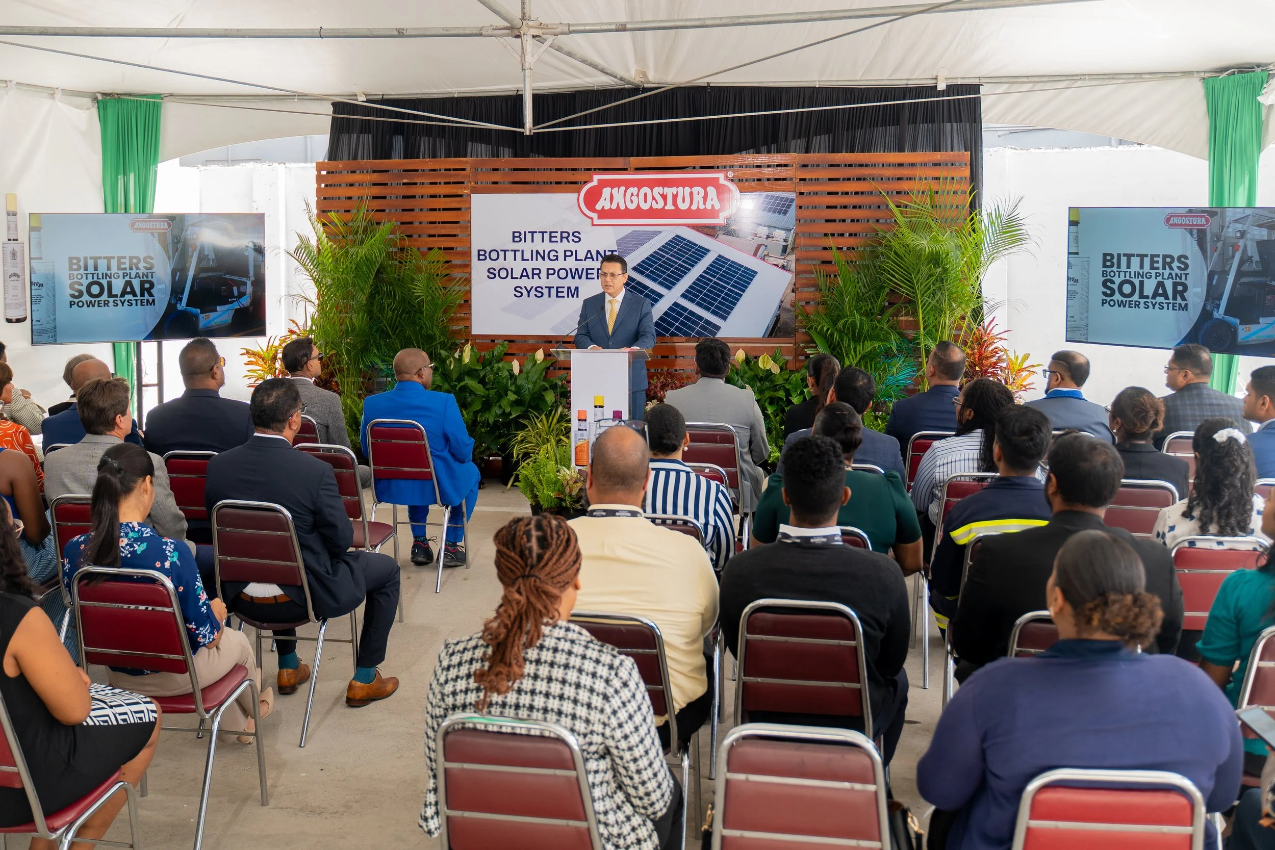 A man in a suit and yellow tie speaking at a presentation event about solar technology. Behind him is a wooden backdrop with a large banner that reads 'Angostura', 'Bitters Bottling Plant Solar Power System', and images of solar panels. The audience is seated in chairs facing the speaker, with screens on either side displaying the same solar power system message. Green plants are in front of the backdrop, and the event is inside a tent.