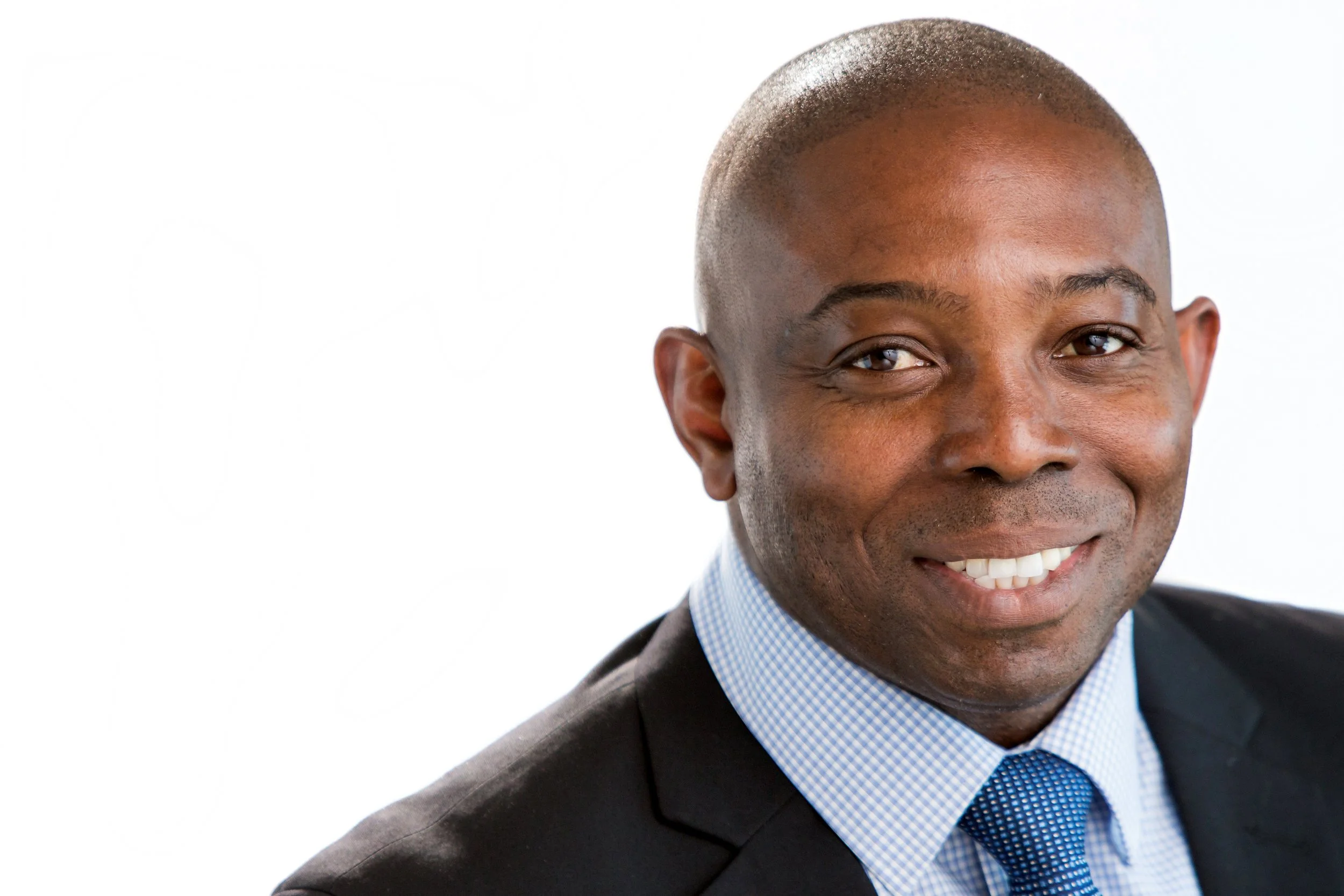 Close-up of a smiling African American man in a dark suit, light blue shirt, and blue tie, against a plain white background.